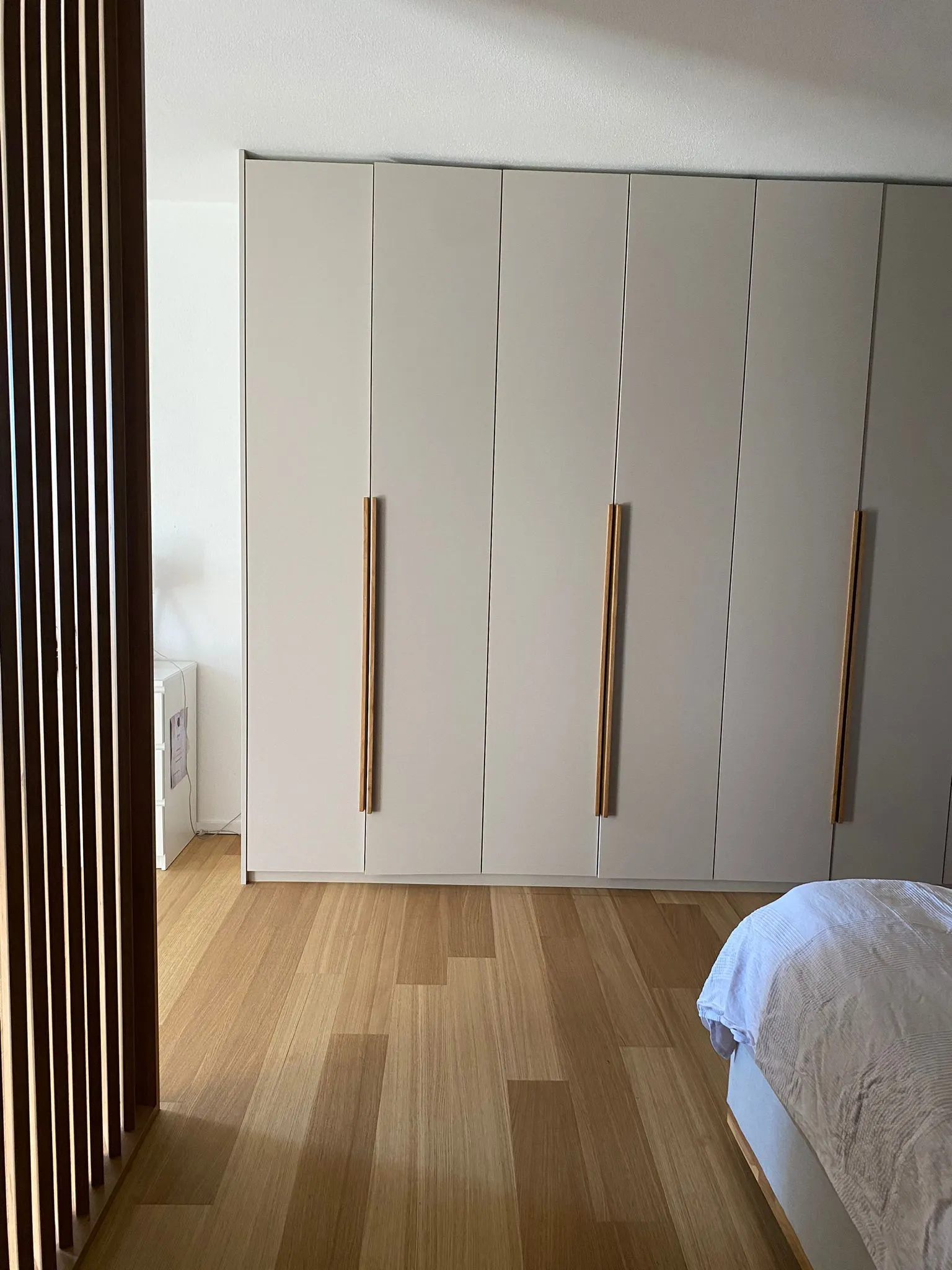 Schlafzimmer mit hellem Holzfußboden, großen grauen Kleiderschränken mit langen Holzgriffen und einem Bett mit weißer Bettwäsche.