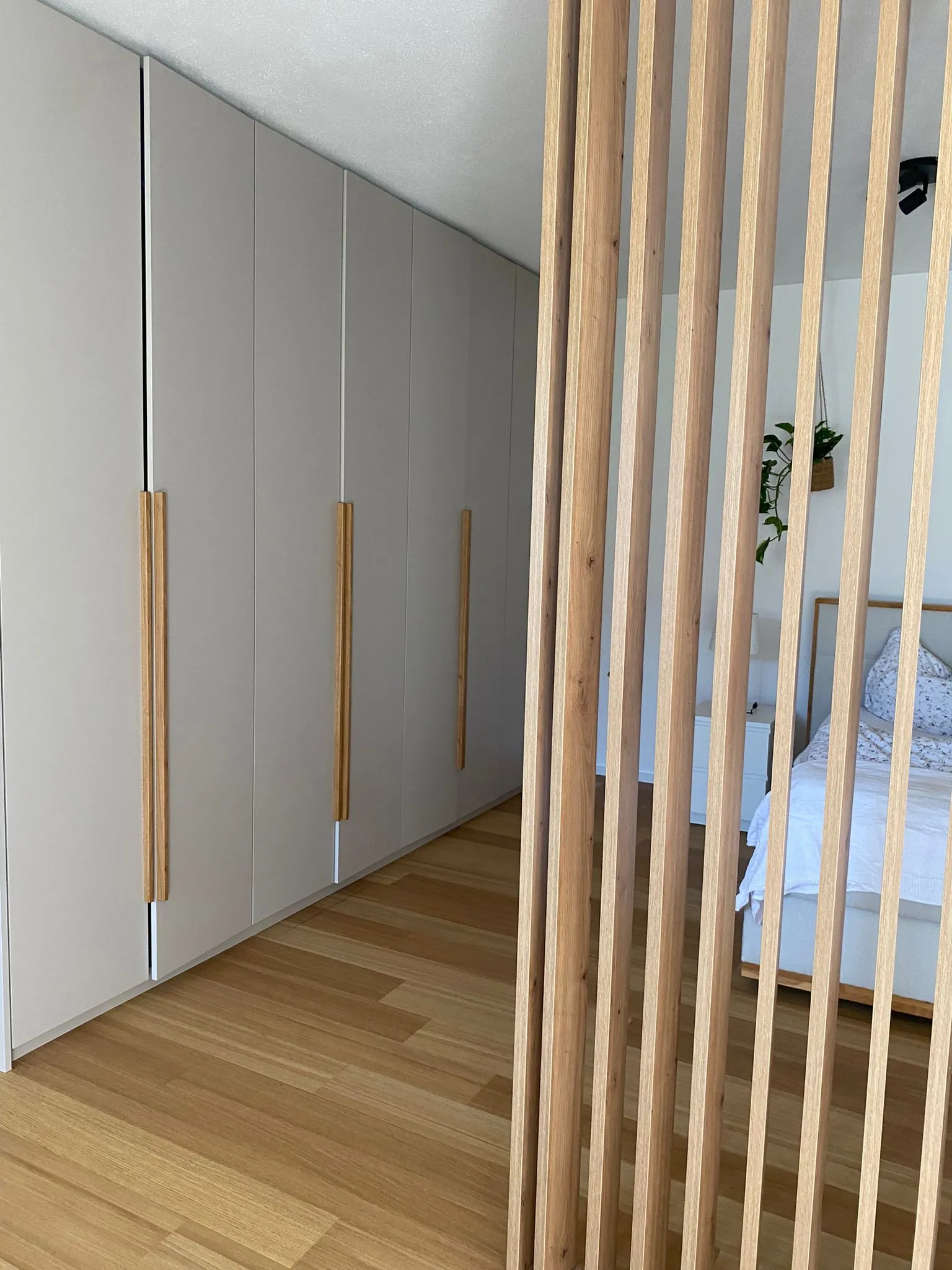 Schlafzimmer mit weißen Schranktüren und langen Holzgriffen neben einem Holzlattenraumteiler, Holzboden und Teil eines Bettes mit weißer Bettwäsche.