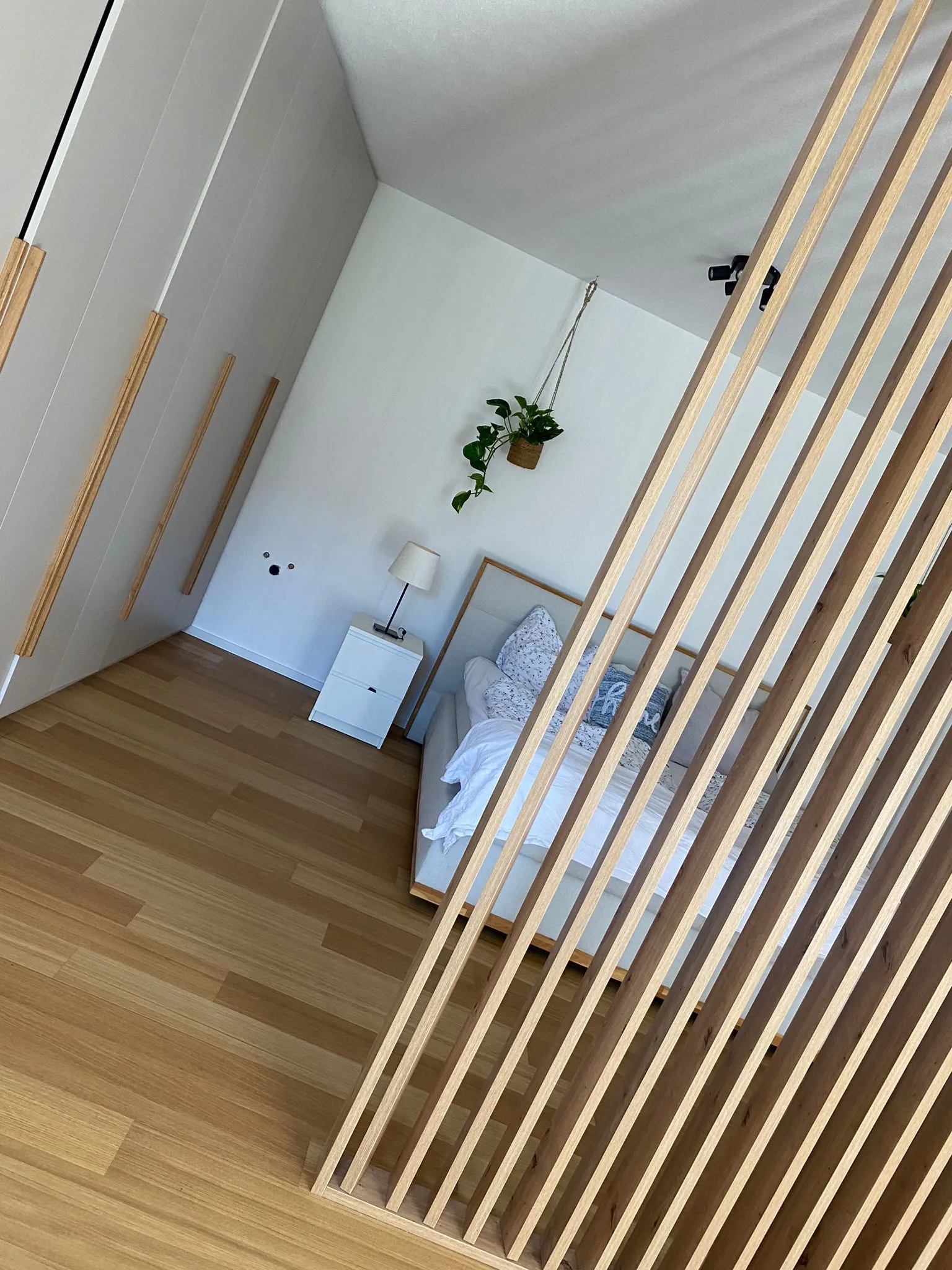 Modernes Schlafzimmer mit Holzlatten als Raumteiler, einem Bett mit weißer Bettwäsche, einem Nachttisch mit Lampe und hängender Pflanze an der weißen Wand.