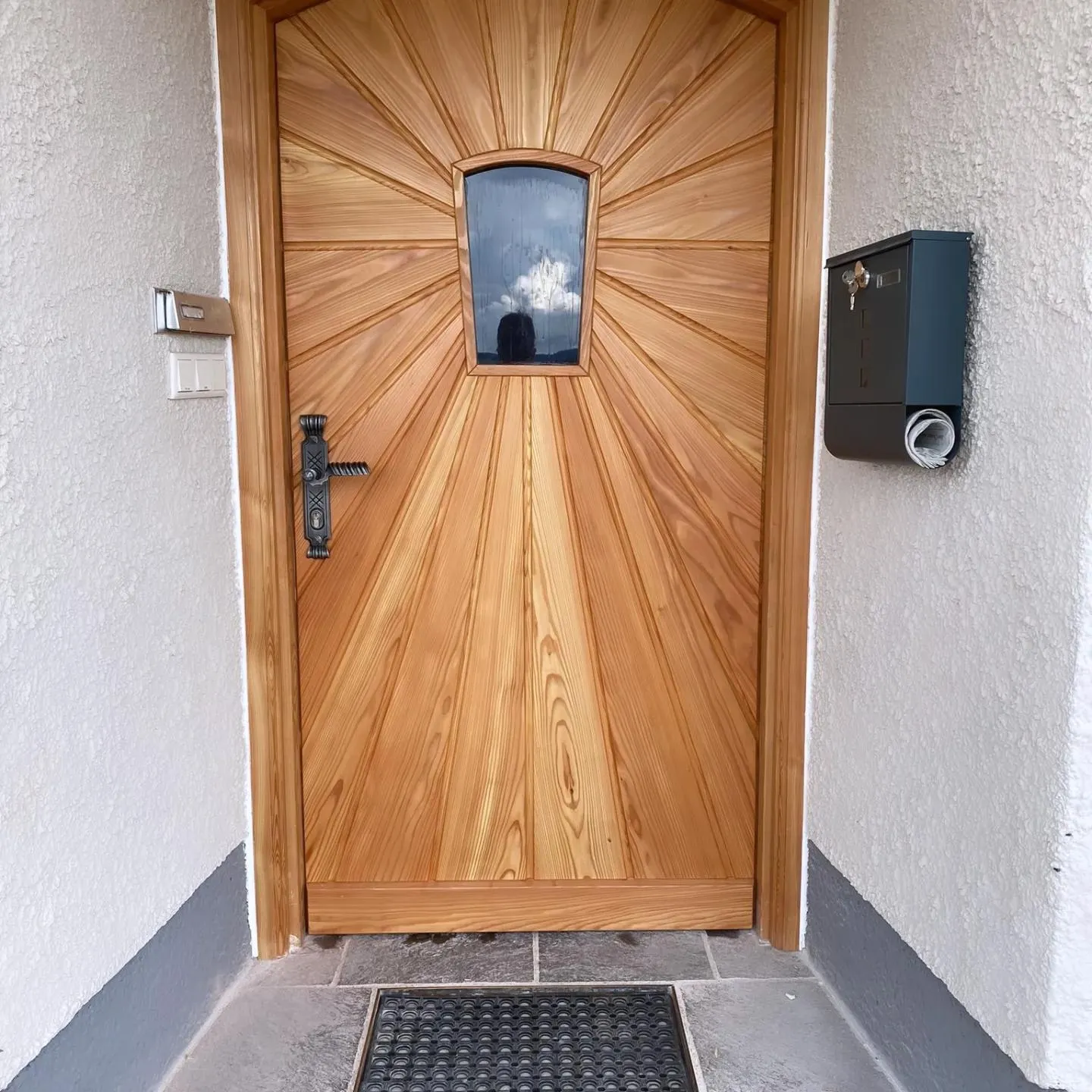 Eingangstür aus Holz mit einem kleinen Fenster und einem schwarzen Türgriff, neben der Tür ein schwarzer Briefkasten mit einem eingesteckten Papier.