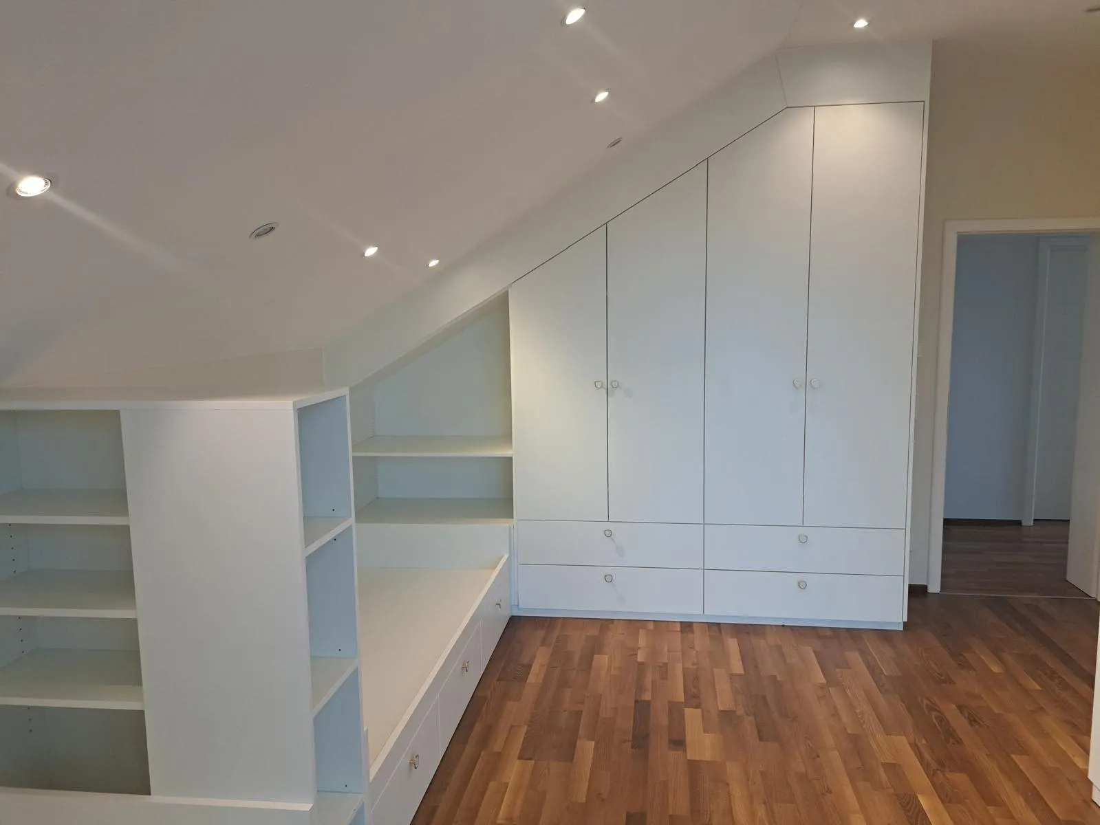 Weißer Einbauschrank mit Schrägdach unter einer Dachschräge und Holzboden in einem hellen Zimmer.