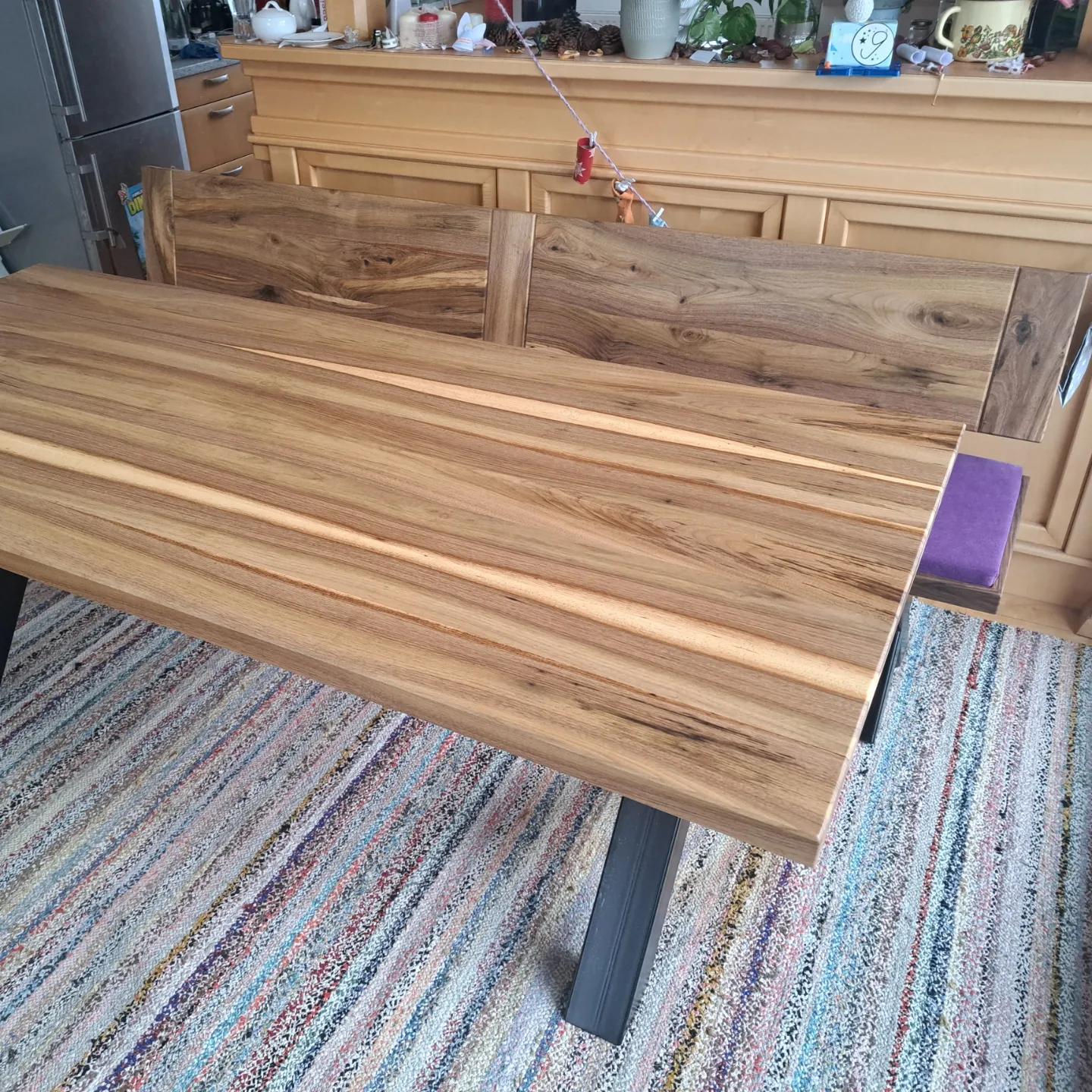 Esstisch und Bank aus hellem Holz mit sichtbarer Maserung auf einem mehrfarbigen Teppich vor einer Kommode mit Dekoration.