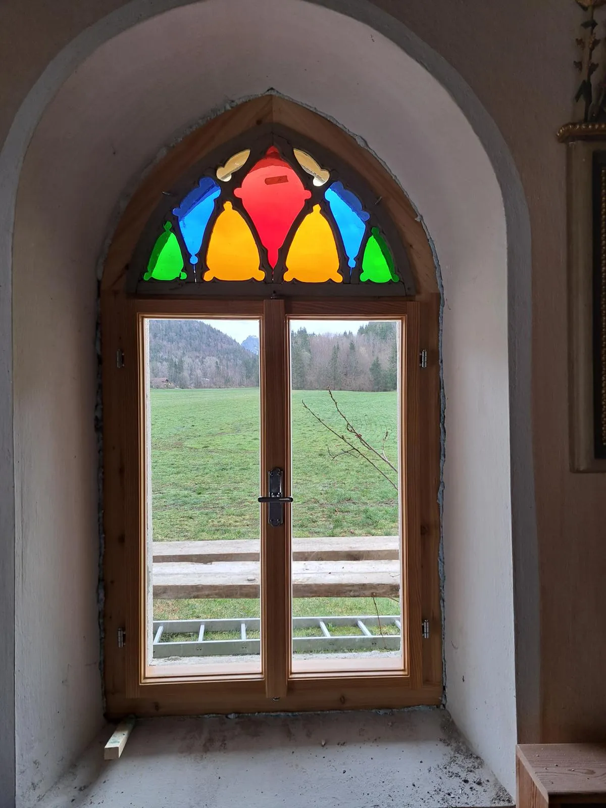 Bunt verglastes Spitzbogenfenster mit Holzrahmen, Blick auf eine Wiese und bewaldete Berge im Hintergrund.