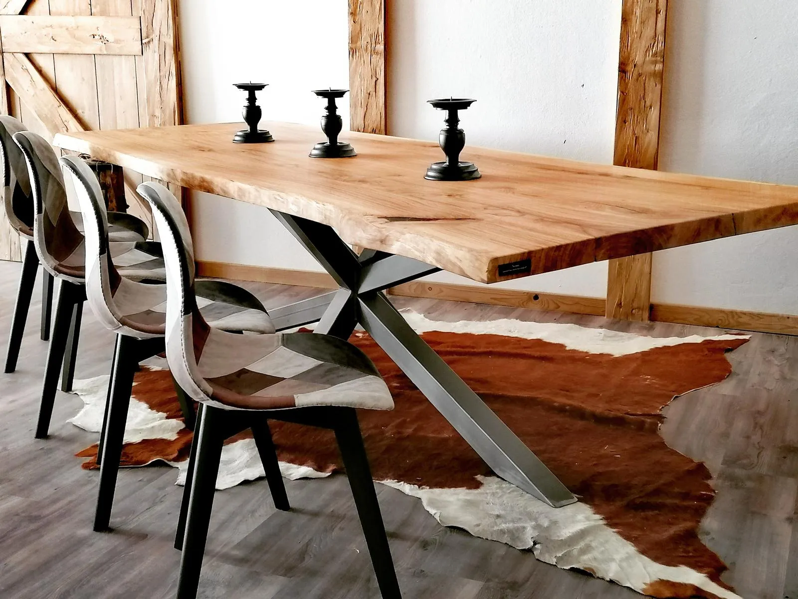 Moderner Esstisch mit einer dicken Holzplatte, Metallgestell und drei gepolsterten Stühlen mit Patchwork-Bezug, darüber drei schwarze Kerzenhalter, auf einem braun-weißen Kuhfellteppich.