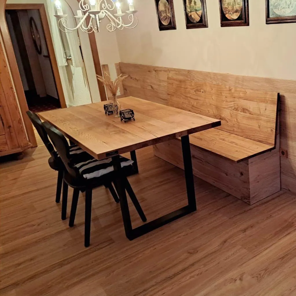 Essbereich mit einem Holztisch, zwei schwarzen Stühlen mit Sitzkissen und einer Holzbank vor einem Wandpaneel mit vier gerahmten Bildern.