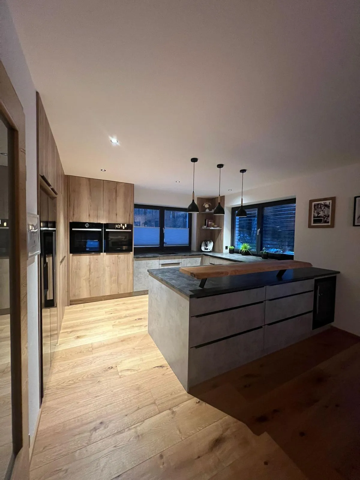 Moderne Küche mit hölzernen Schränken, zentraler Insel aus Betonoptik, harten Holzböden und großen Fenstern mit Innenjalousien.