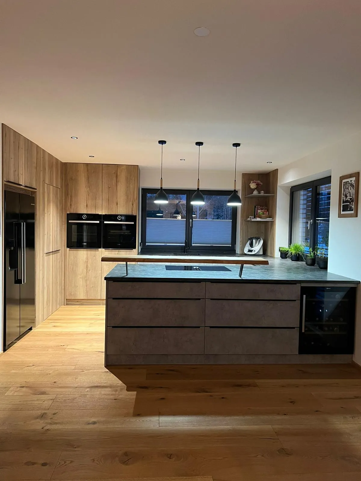 Moderne Küche mit Holzfußboden, grauer Kücheninsel mit Herd, drei hängenden Lampen, eingebauten Backöfen und großen Fenstern.