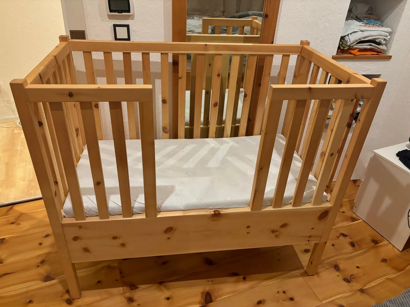 Holz-Kinderbett mit weißen Matratze in einem Zimmer mit Holzfußboden und Spiegel an der Wand.