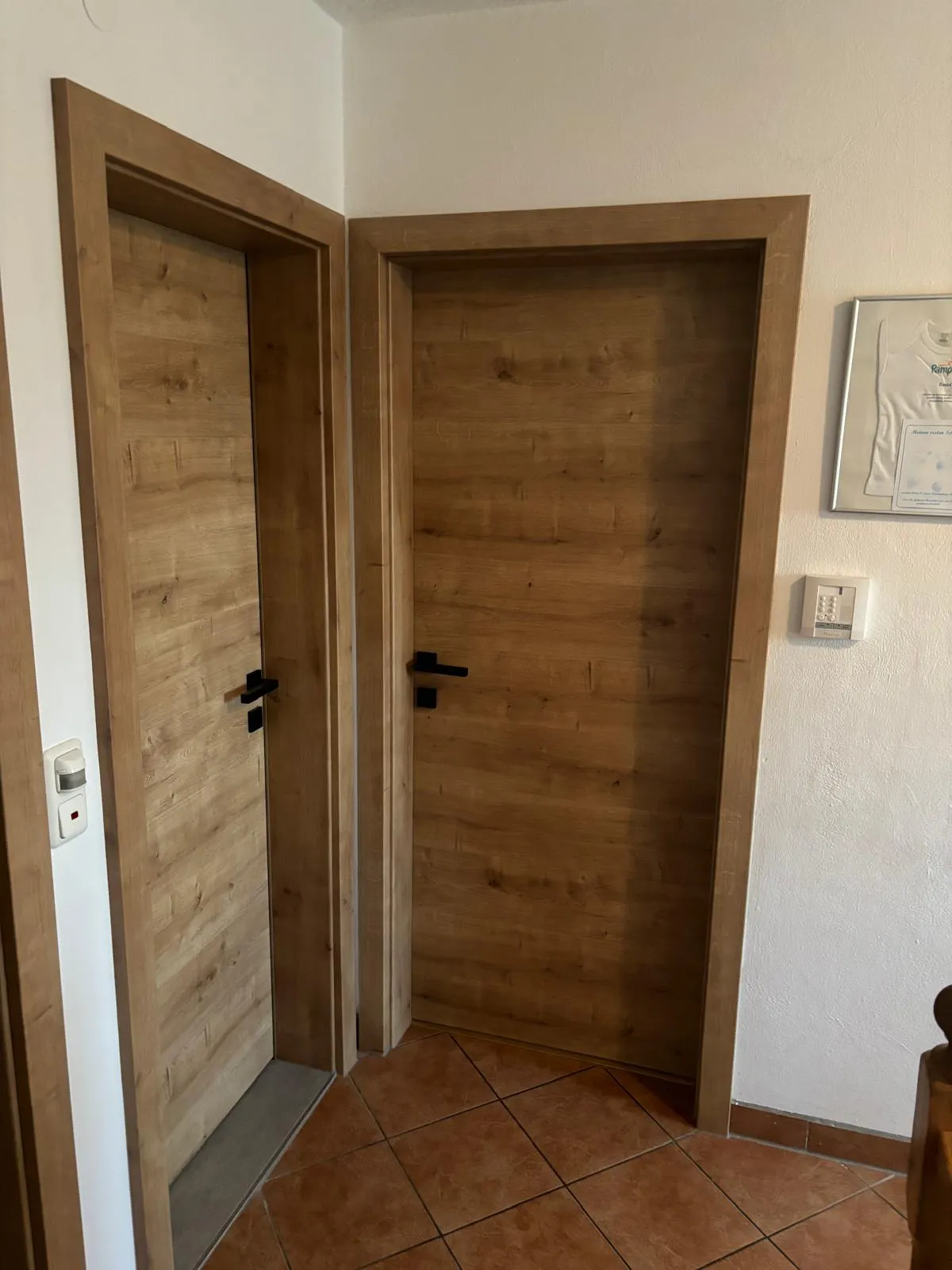 Zwei geschlossene Holztüren mit schwarzen Griffen in einem Flur mit braunen Fliesenboden und weißen Wänden.