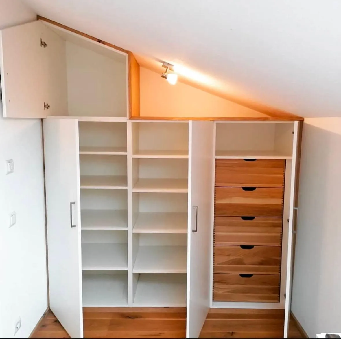 Weiße Einbauschränke mit offenen Regalen und Holzziehschubladen unter einer Schräge mit Deckenleuchte.