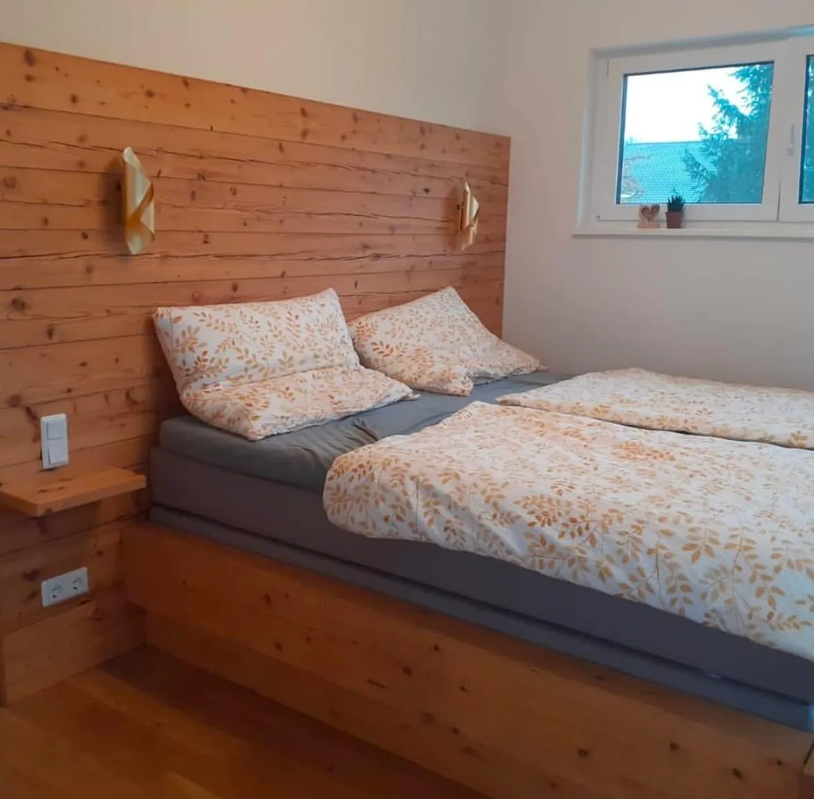 Gemütliches Schlafzimmer mit großem Holzbett, weiß-gelben Bettdecken mit Blattmuster und Fenster mit Ausblick auf Bäume.