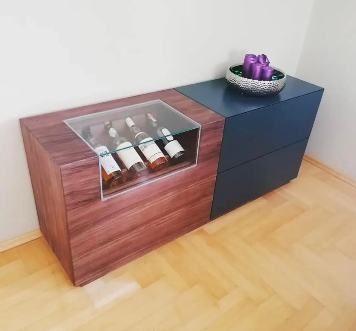 Moderner Schrank mit Holz- und blauen Schubladenanteilen, seitlich Fach mit Weinflaschen unter Glas und silberner Schale mit violetten Kerzen oben.