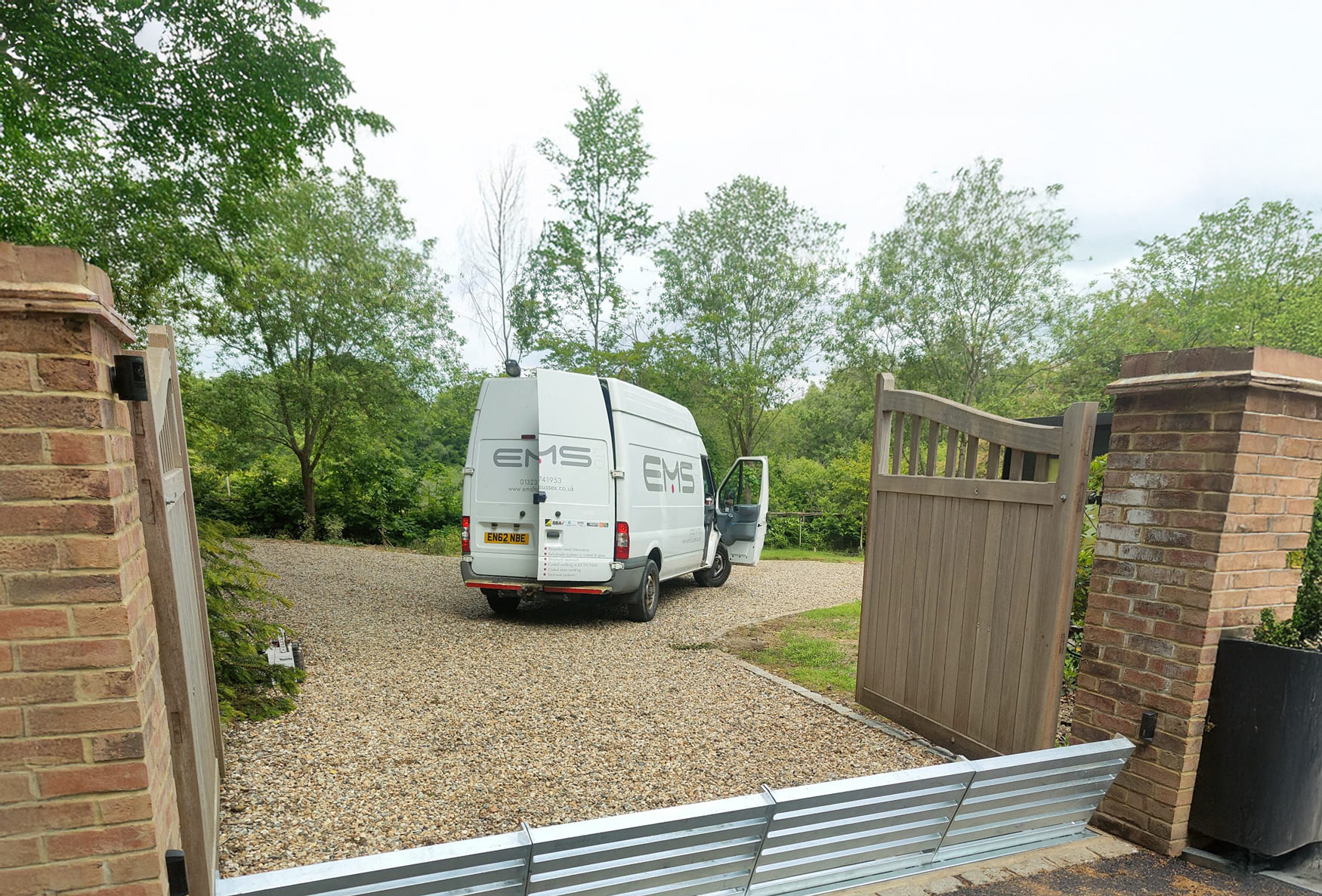 A delivery van with open back doors is parked on a gravel driveway between brick pillars.