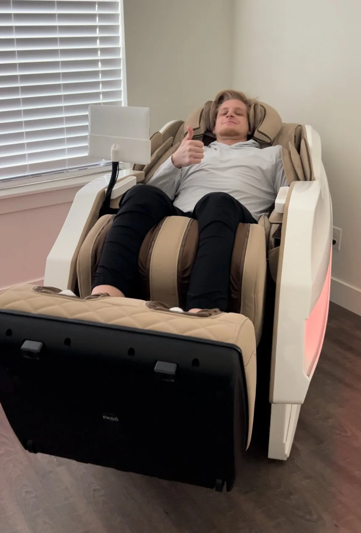 Mies rentoutuu hierontatuolissa, peukalo pystyssä, vaaleanharmaassa paidassa ja mustissa housuissa, tuoli on beige ja valkoinen, ikkuna taustalla.