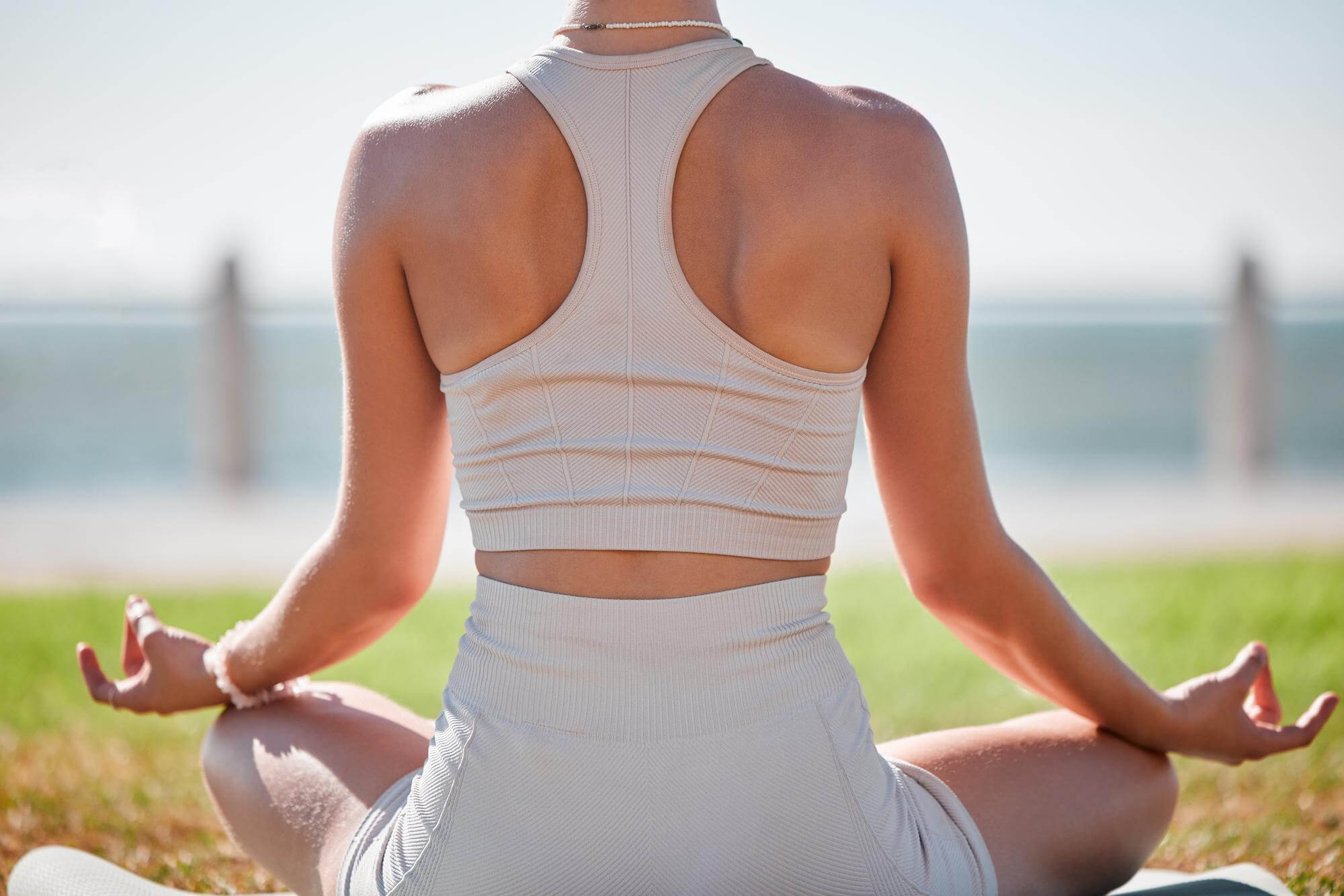 Nainen joogaamassa ulkona, yllään urheiluvaatteet, taustalla meri ja vihreä nurmikko, valoisa ja aurinkoinen sää.