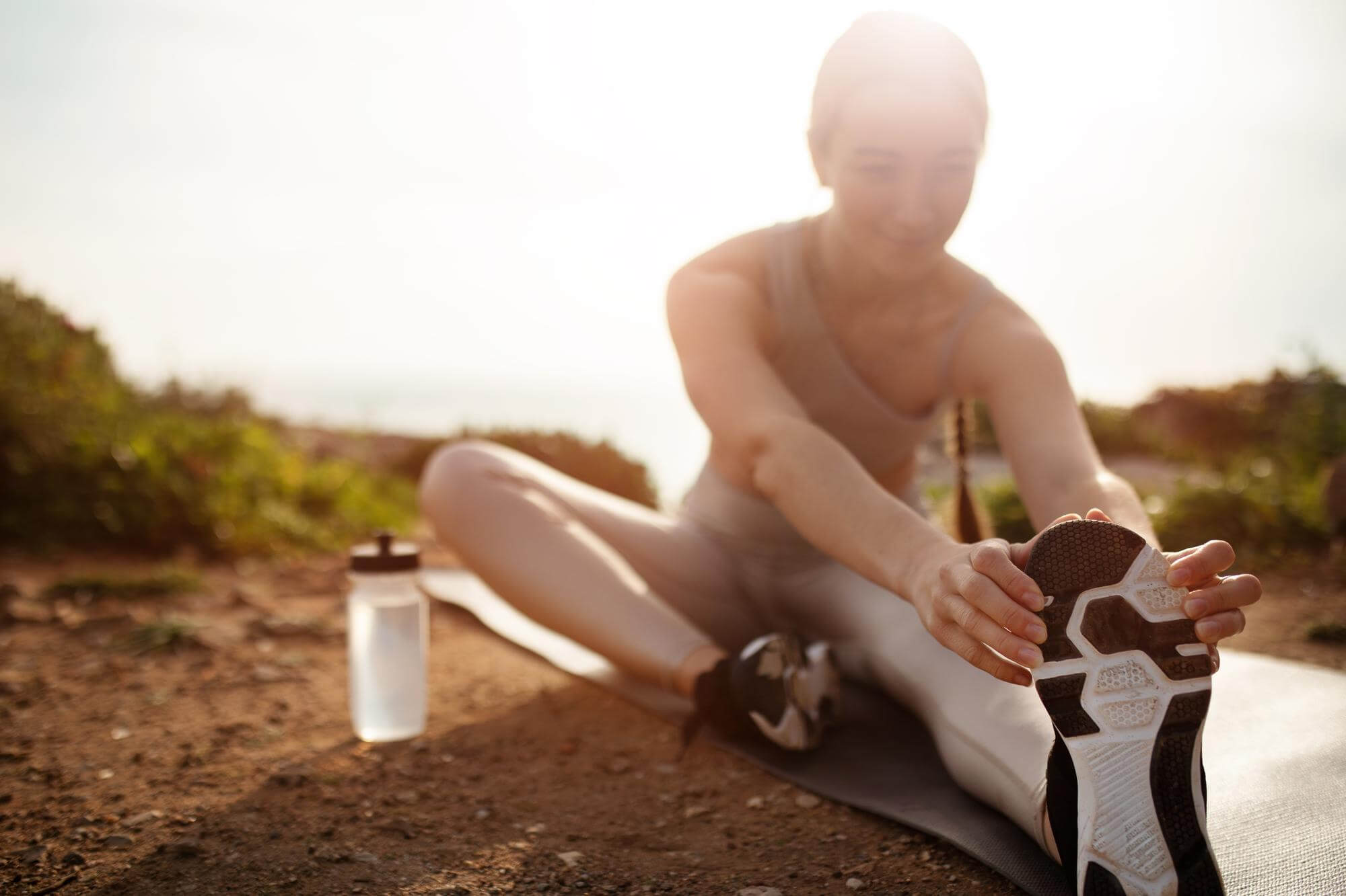Nainen venyttelee ulkona joogamatolla, juomapullo vieressä, aurinkoisessa säässä.