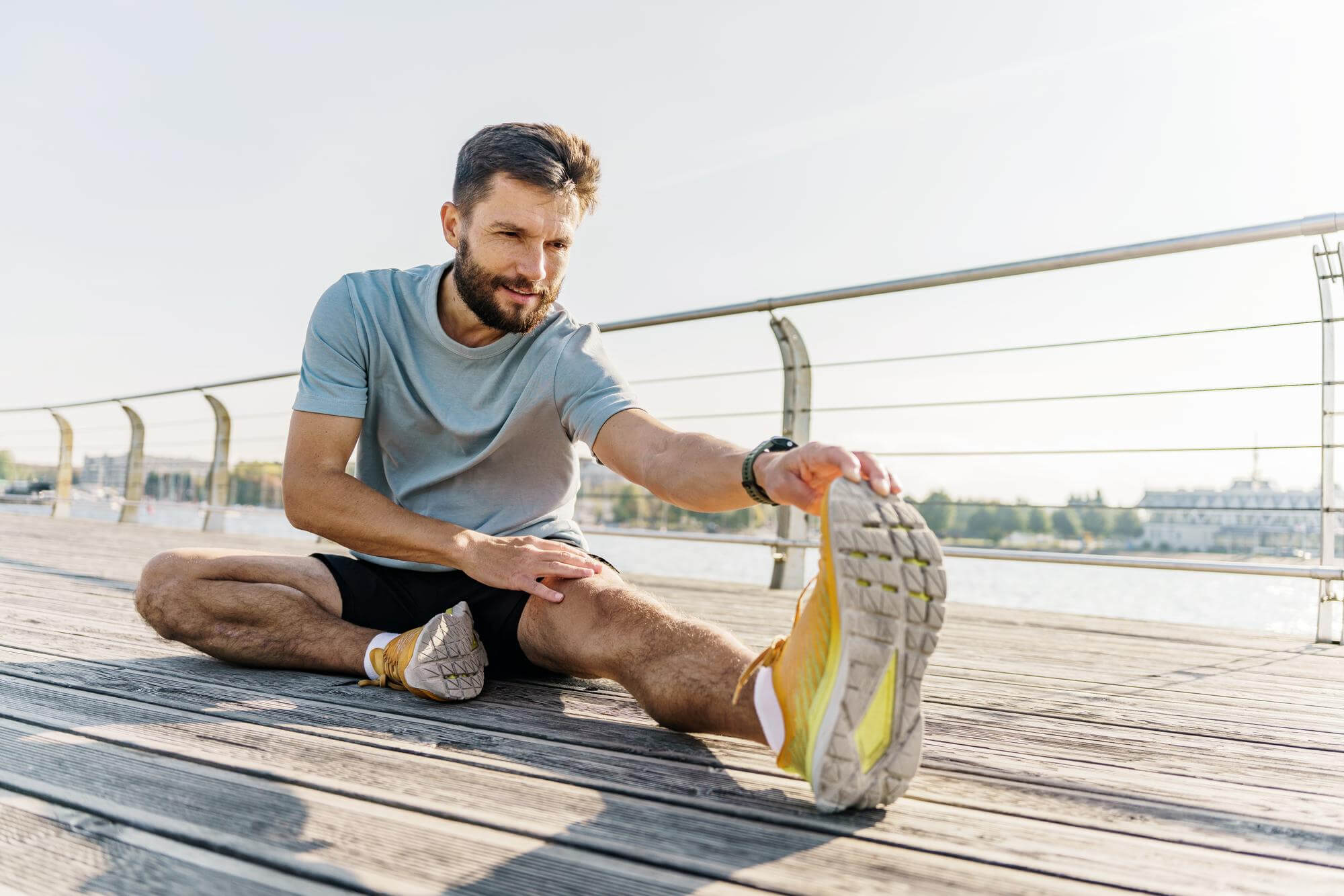 Keskittynyt mies venyttelee jalkojaan ulkona, aurinkoisessa säässä, puulattialla, taustalla vesi ja rakennuksia. Hänellä on harmaa paita ja mustat shortsit.