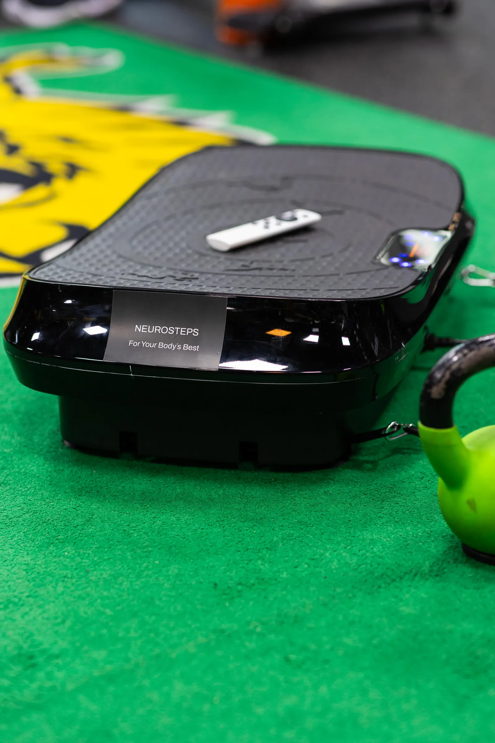 Kuvassa on Neurosteps-värähtelylaite, jossa on kaukosäädin päällä, vihreällä alustalla ja vihreä kahvakuula vieressä. Laitteessa on musta runko ja harmaa pinta.