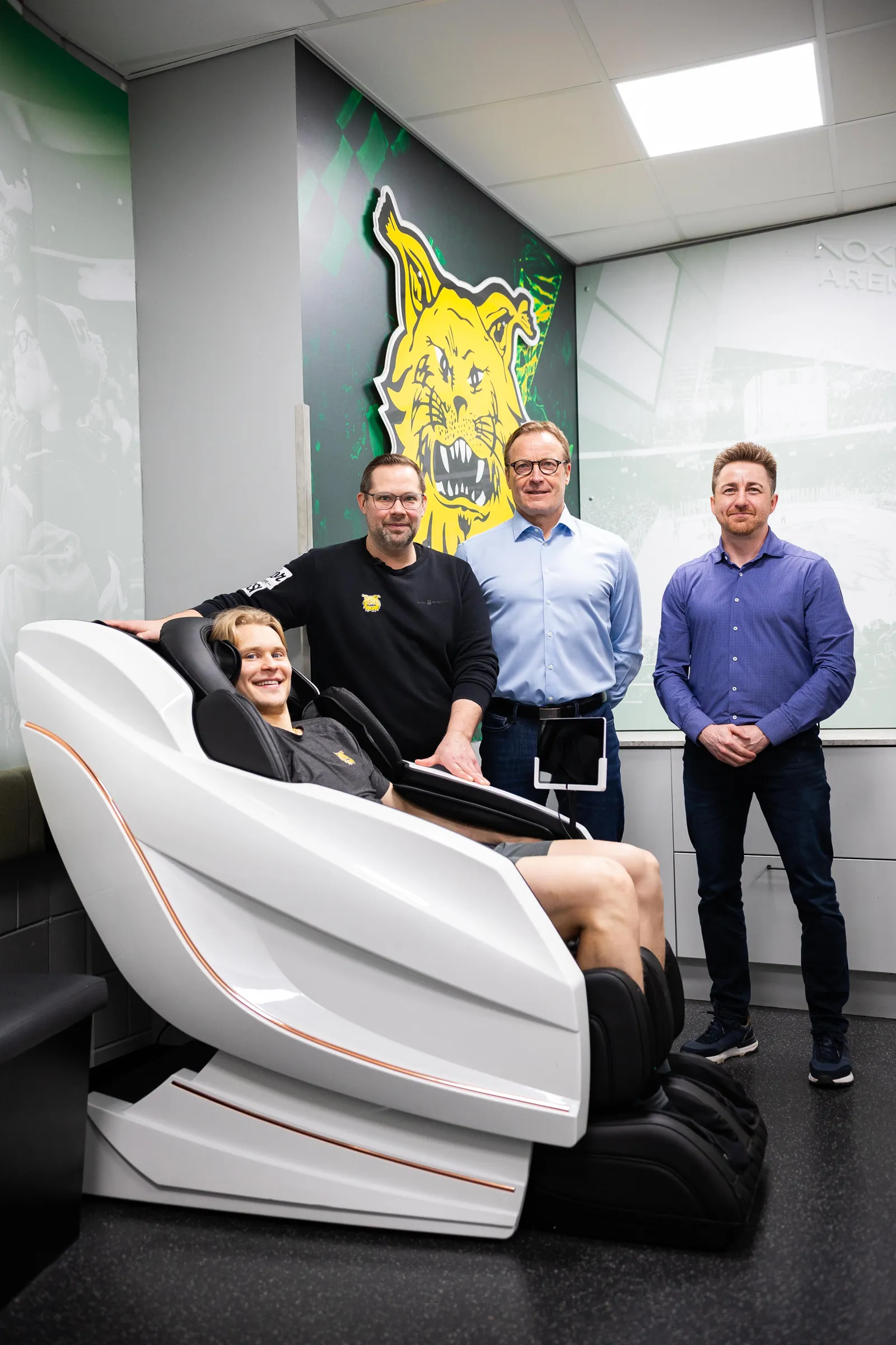 Neljä miestä poseeraa hierontatuolin kanssa huoneessa, jossa on joukkueen logo ja Nokia Arenan kuva seinällä. Mies istuu tuolissa hymyillen, kun taas muut seisovat hänen takanaan.