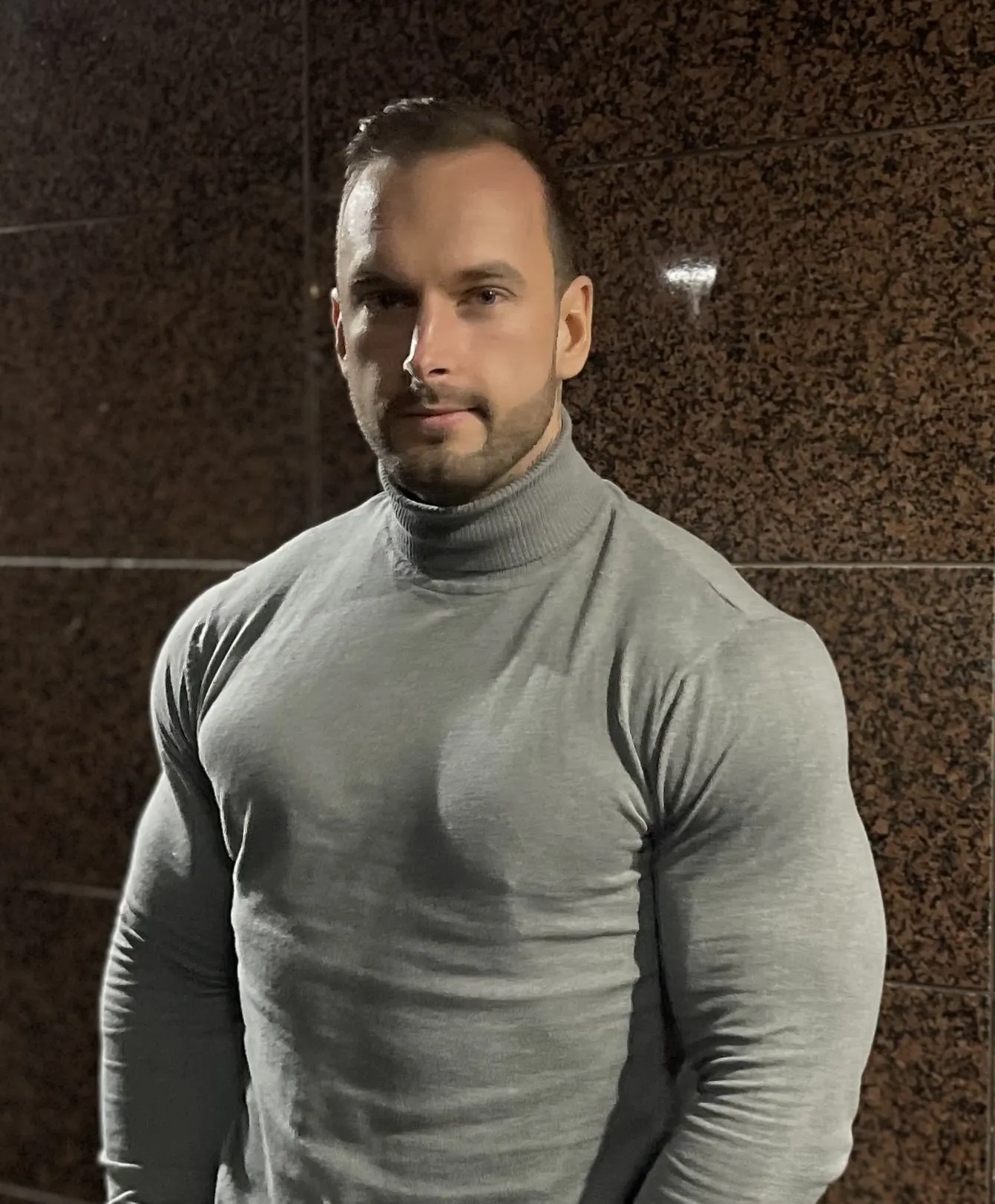 Lihaksikas mies harmaassa poolopaidassa seisoo ruskean seinän edessä, valaistus on pehmeä ja korostaa hänen lihaksiaan.