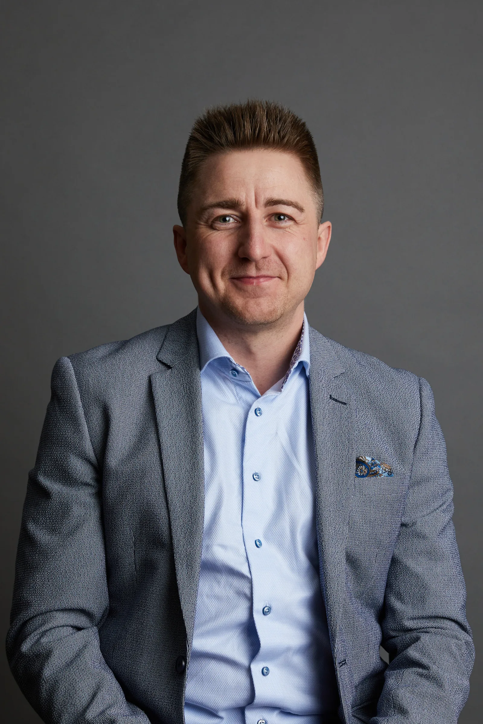 Muotokuva hymyilevästä miehestä vaaleansinisessä paidassa ja harmaassa bleiserissä, jossa on taskuliina harmaata taustaa vasten.