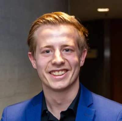 Jonge man met blond haar en blauwe jas glimlacht op een donkere achtergrond.