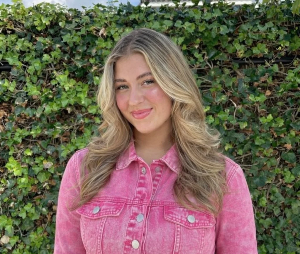 Vrouw met blonde krullende haren draagt een roze spijkerjack en poseert voor een bladvormige groene haag.