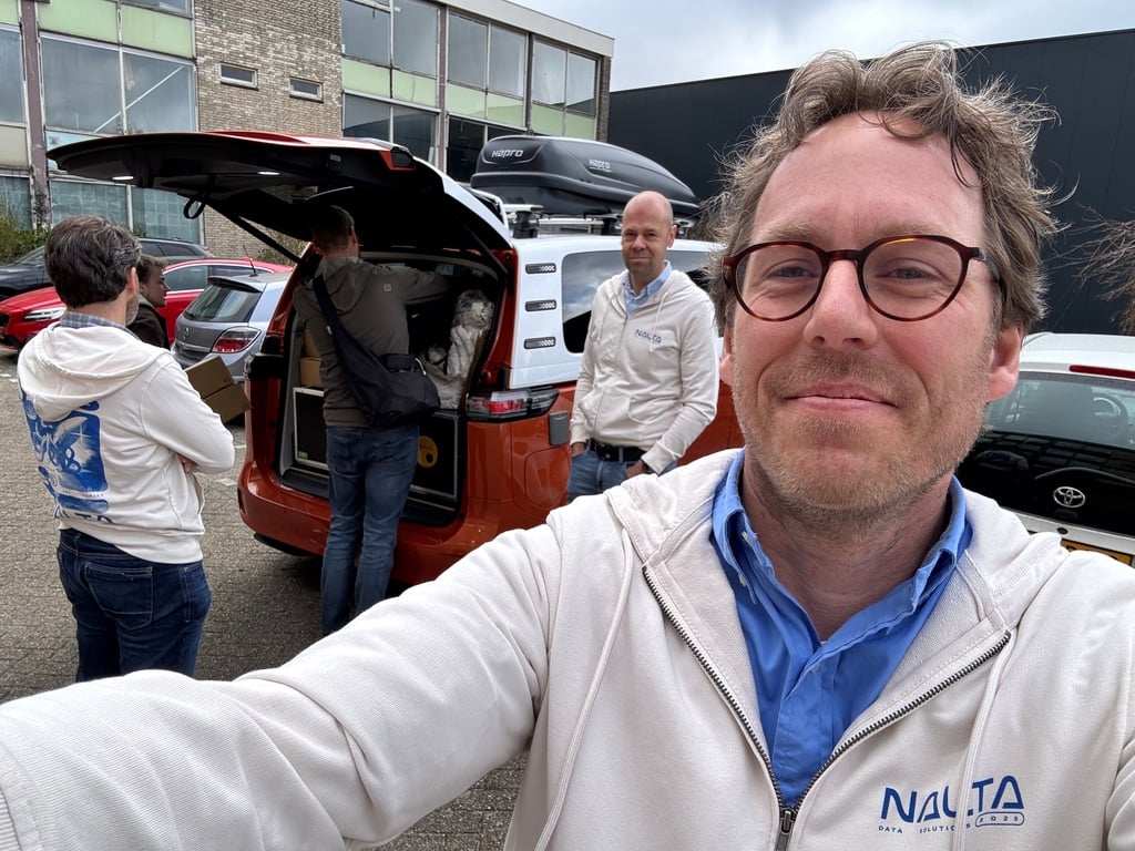 Vier mannen met Nalta-hoodies bij een auto met open kofferbak vol uitrusting, één maakt selfie.