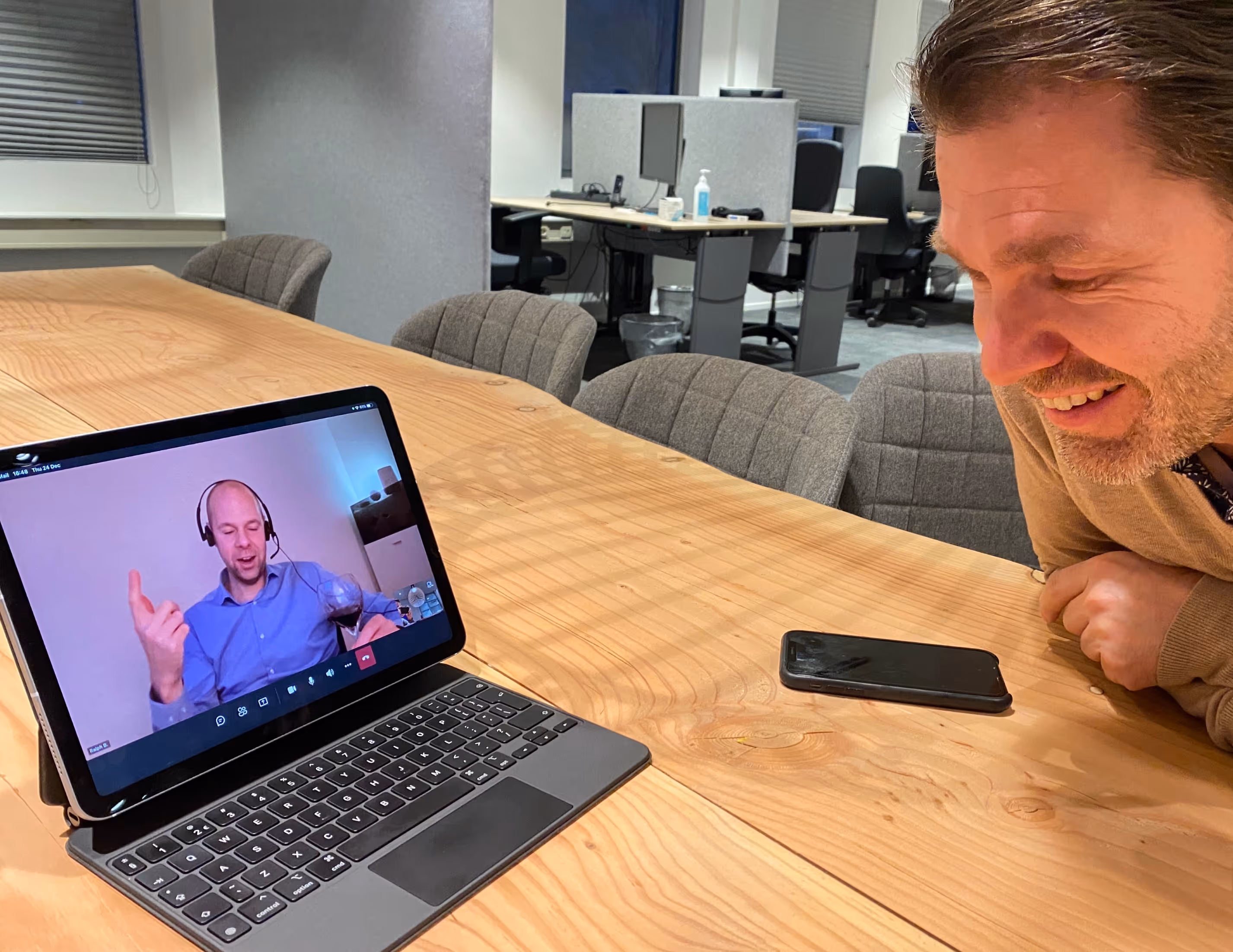 Man lacht naar tablet met videovergadering waar een man met headset wijn vasthoudt.