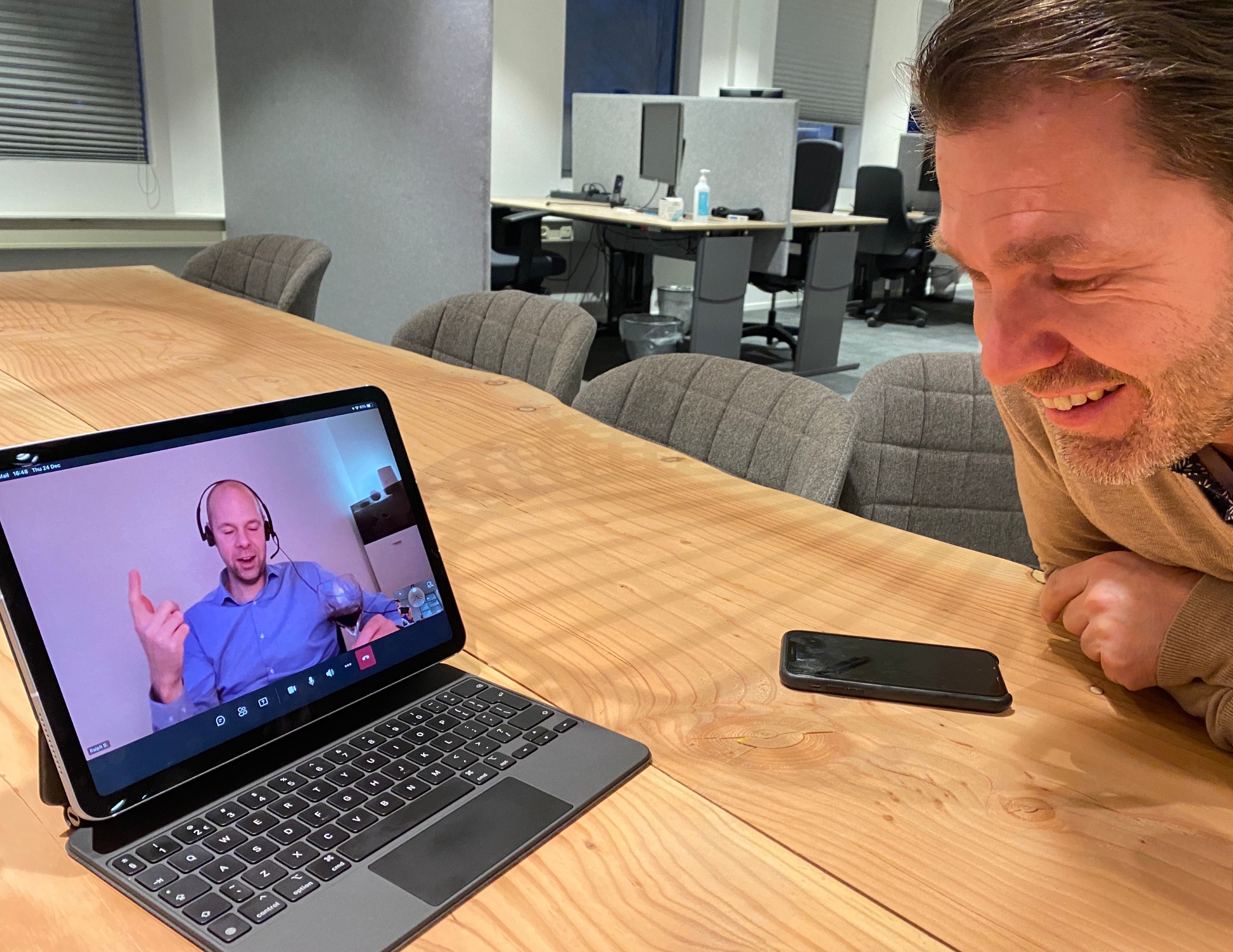 Man lacht naar tablet met videovergadering waar een man met headset wijn vasthoudt.