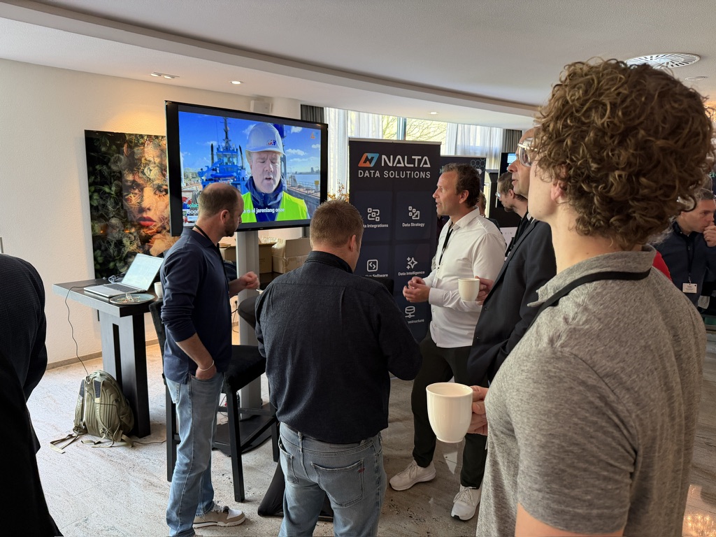 Mannen praten bij een stand met een scherm en banner van Nalta Data Solutions, sommigen met koffie in de hand.