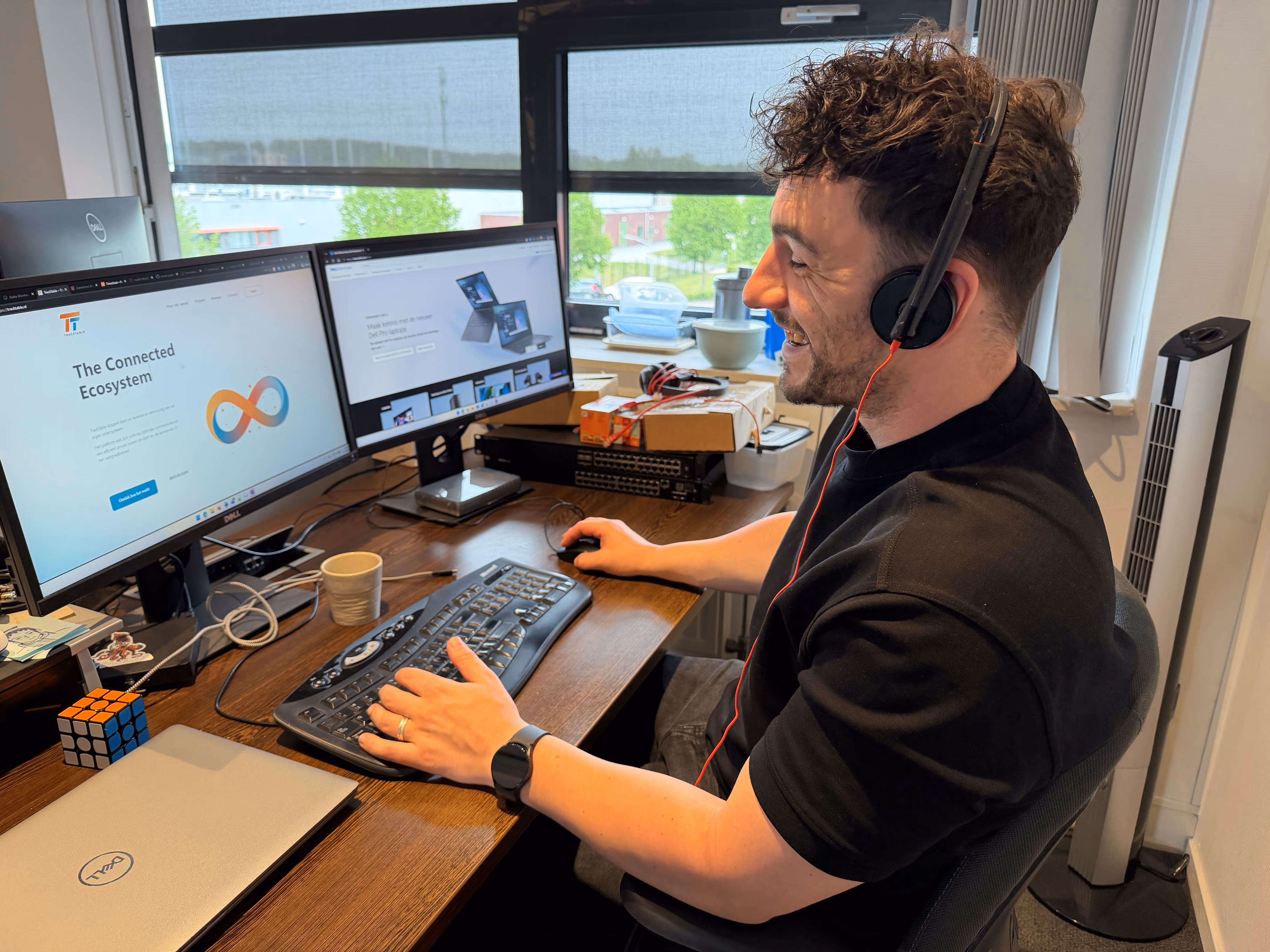 Man met headset aan bureau met twee computerschermen en Rubiks kubus, werkend in kantoor met raam.
