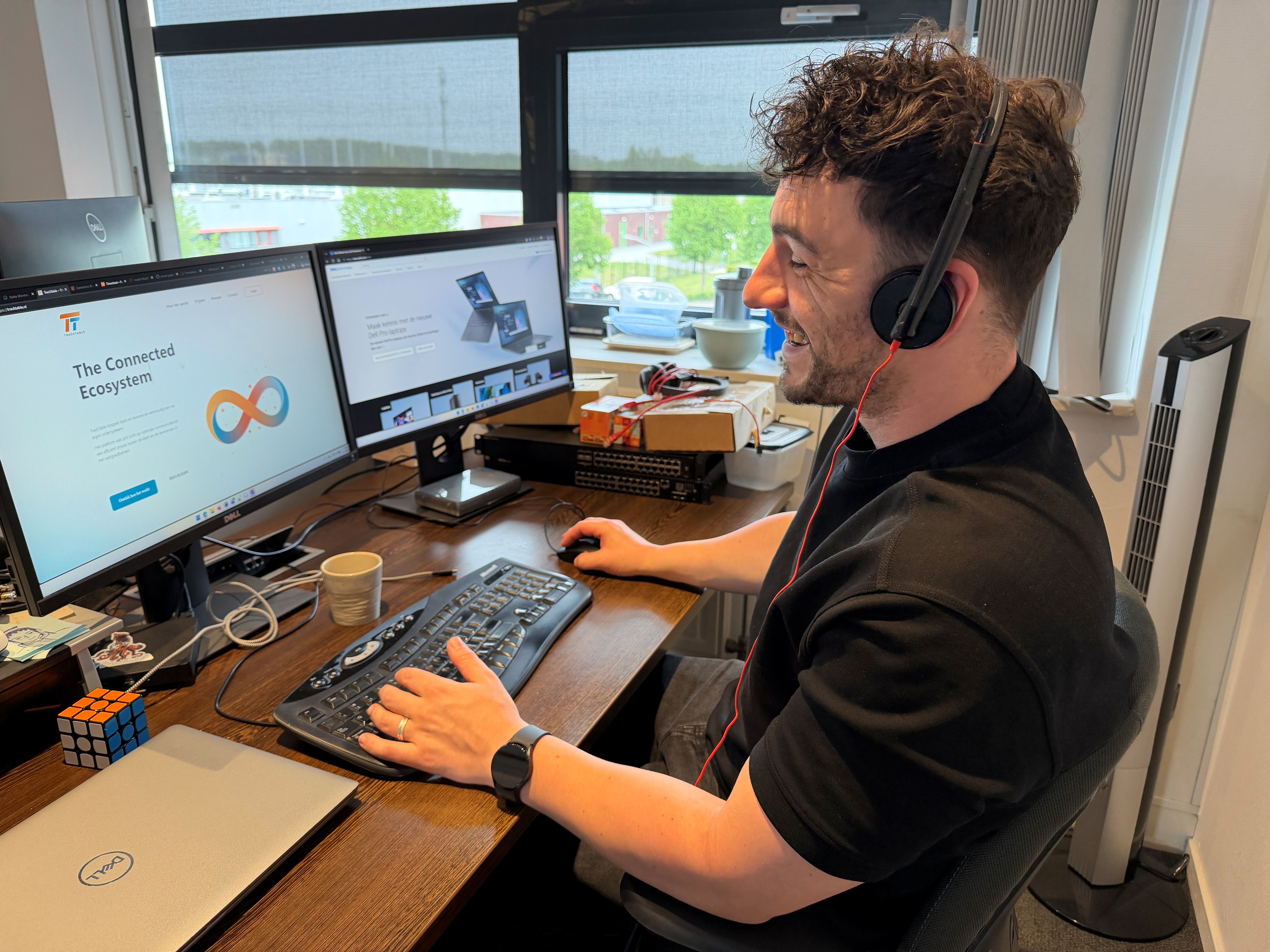 Man met headset aan bureau met twee computerschermen en Rubiks kubus, werkend in kantoor met raam.
