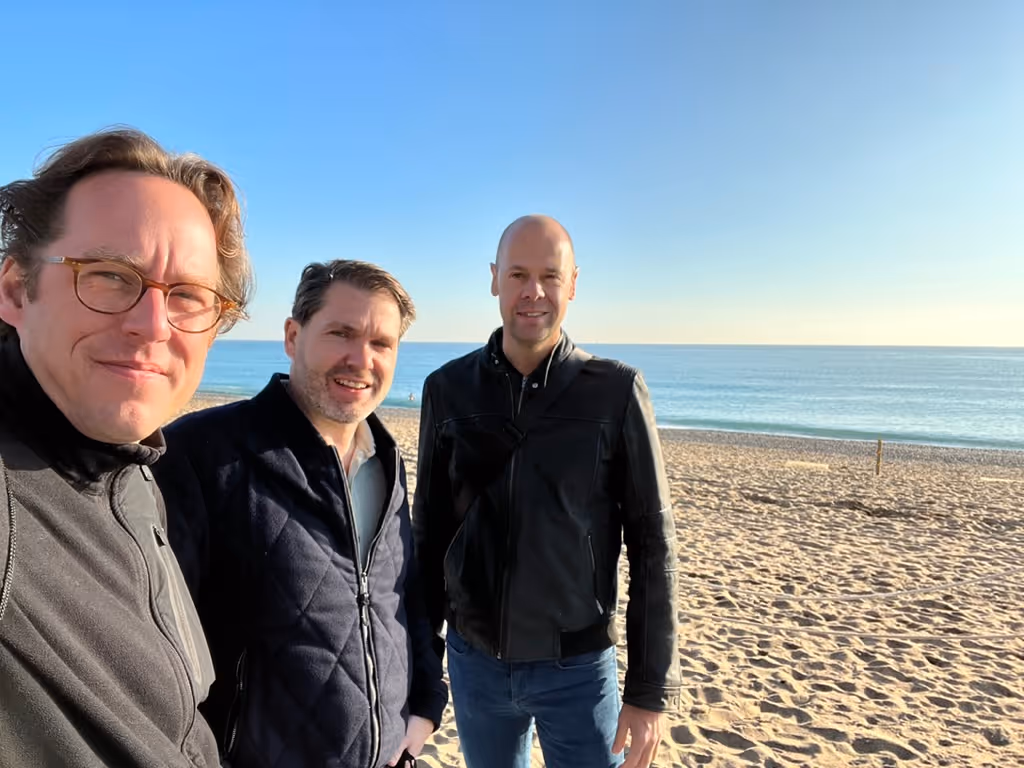 Drie mannen staan op een zonnig strand met blauwe lucht en zee op de achtergrond.