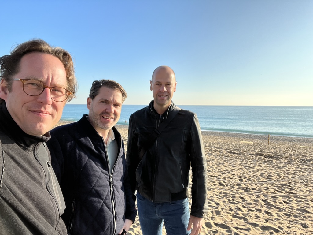 Drie mannen staan op een zonnig strand met blauwe lucht en zee op de achtergrond.