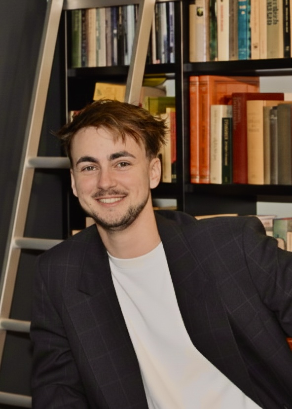 Jonge man met warrig haar en gelaatsbeharing, glimlachend, zittend voor een boekenplank en ladder.