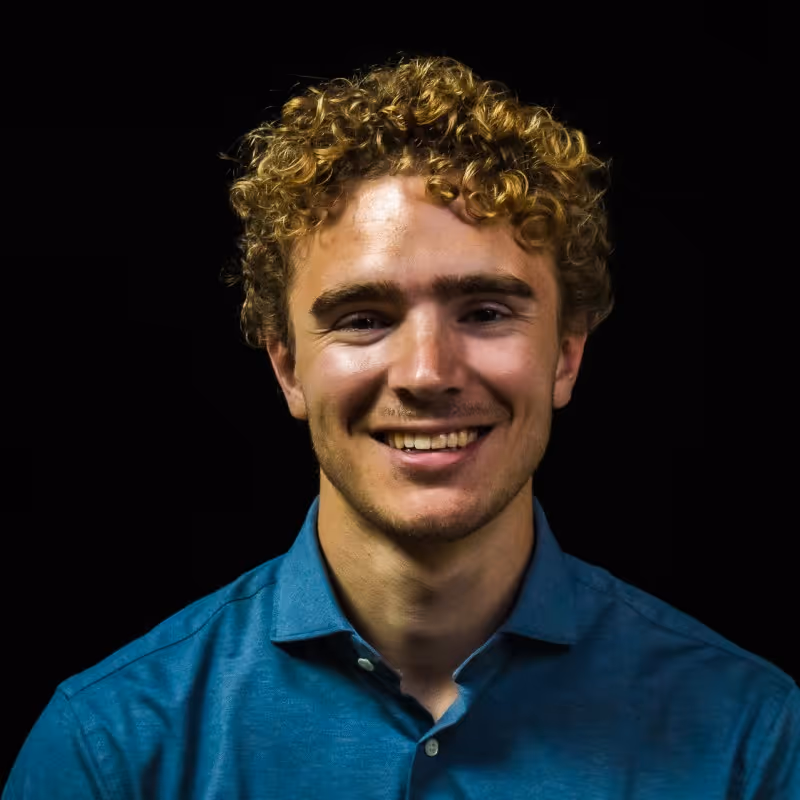 Jonge man met krullend blond haar en blauwe overhemd glimlacht tegen zwarte achtergrond.