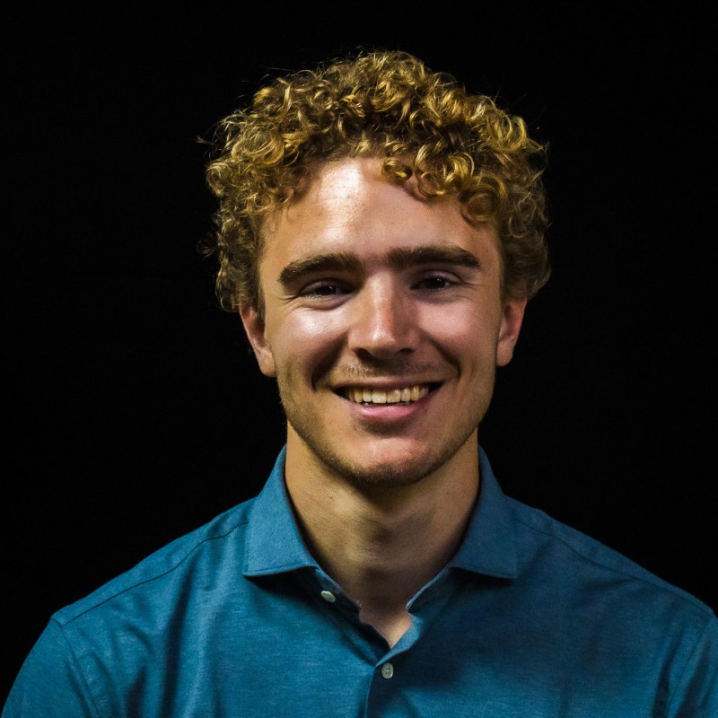 Jonge man met krullend blond haar en blauwe overhemd glimlacht tegen zwarte achtergrond.