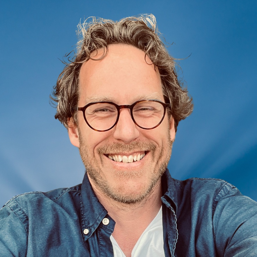 Man met krullend haar en bril glimlacht breed met blauwe lucht op de achtergrond.