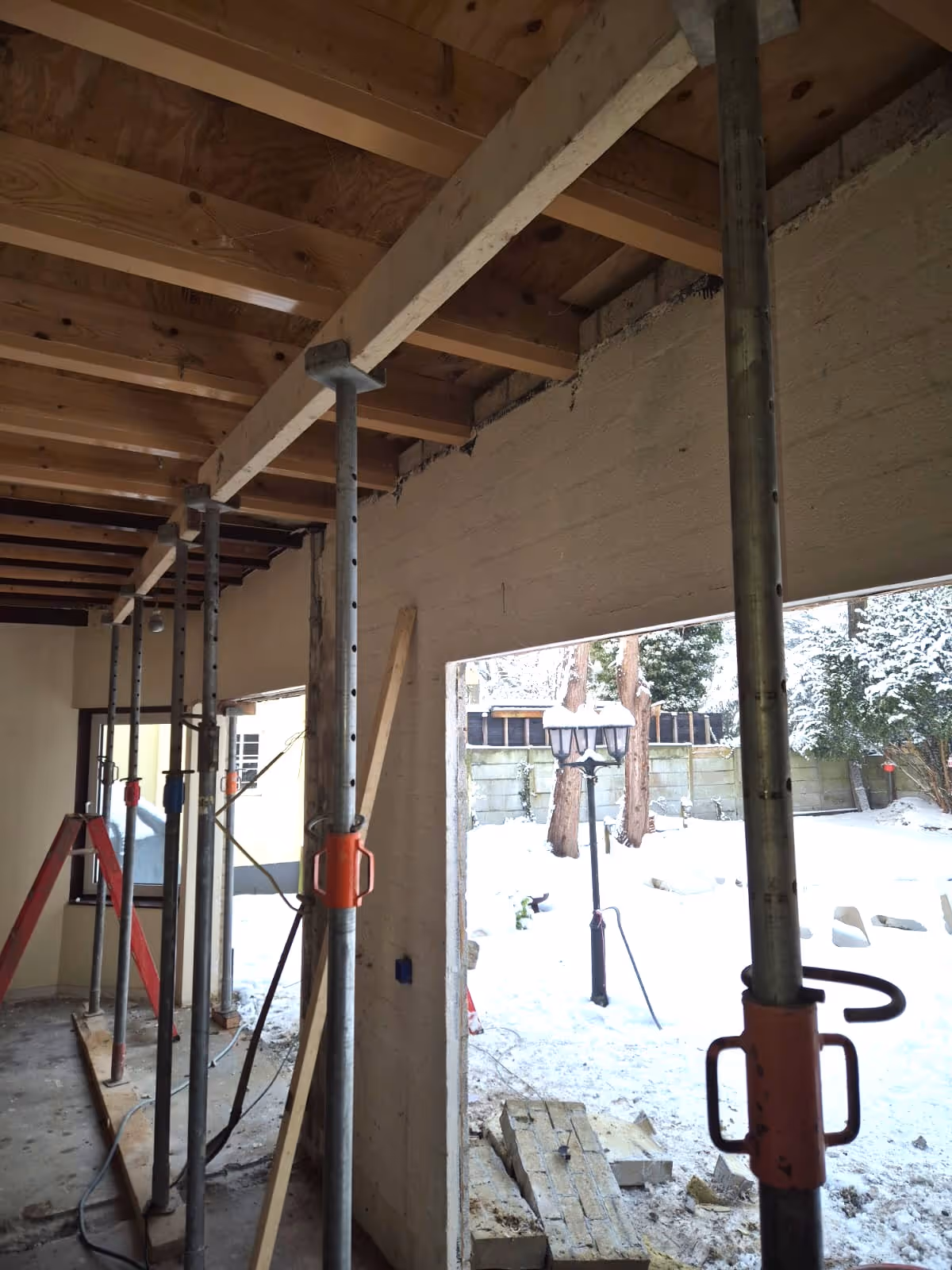 Interior view of a construction site showing metal support poles holding up wooden ceiling beams with an open wall revealing a snowy outdoor yard with trees and a lamp post.