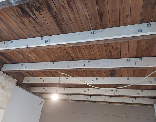 Wooden ceiling beams with metal brackets installed for roof reinforcement, with a hanging light bulb and exposed wiring.