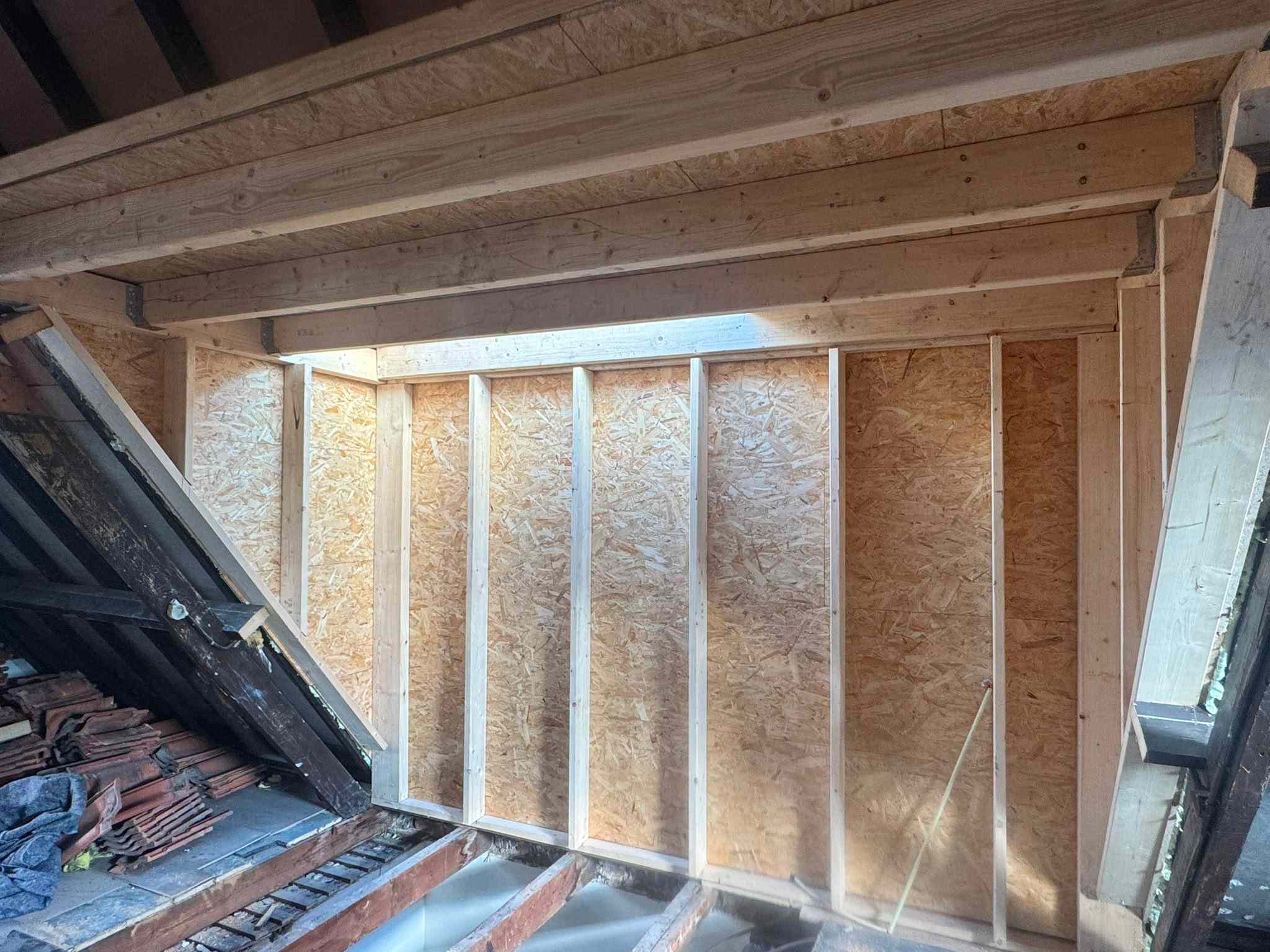 Wooden ceiling beams with metal brackets installed for roof reinforcement, with a hanging light bulb and exposed wiring.