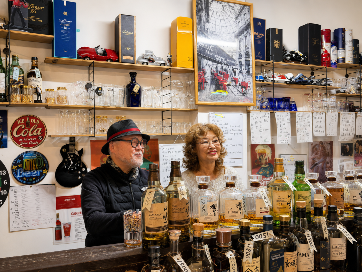 Husband-and-wife owners behind the counter of a small Japanese whisky bar in Osaka, surrounded by bottles of Yamazaki, Hibiki and other Japanese whiskies.