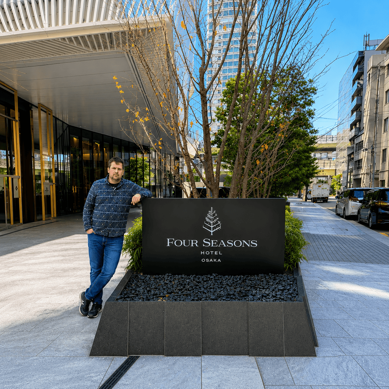 Dylan Clements standing beside the Four Seasons Hotel Osaka entrance sign during his stay in Japan. The modern luxury hotel entrance features contemporary architecture, landscaped gardens, and the iconic Four Seasons logo, with Osaka's city skyline in the 