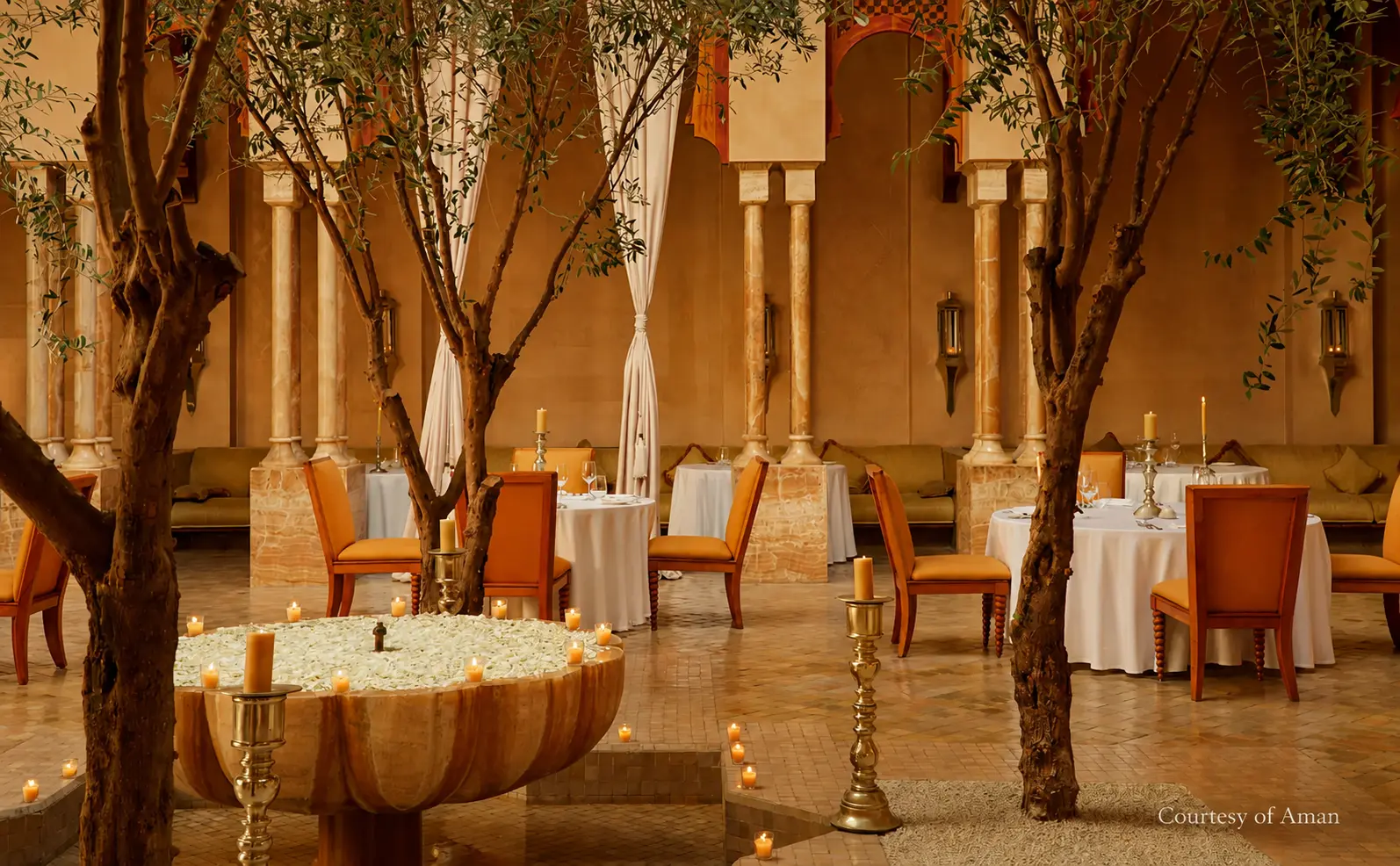 The courtyard dining room at The Moroccan Restaurant at Amanjena in Marrakech, featuring olive trees, candlelit tables, traditional Moroccan arches, and elegant architecture. Image courtesy of Aman.