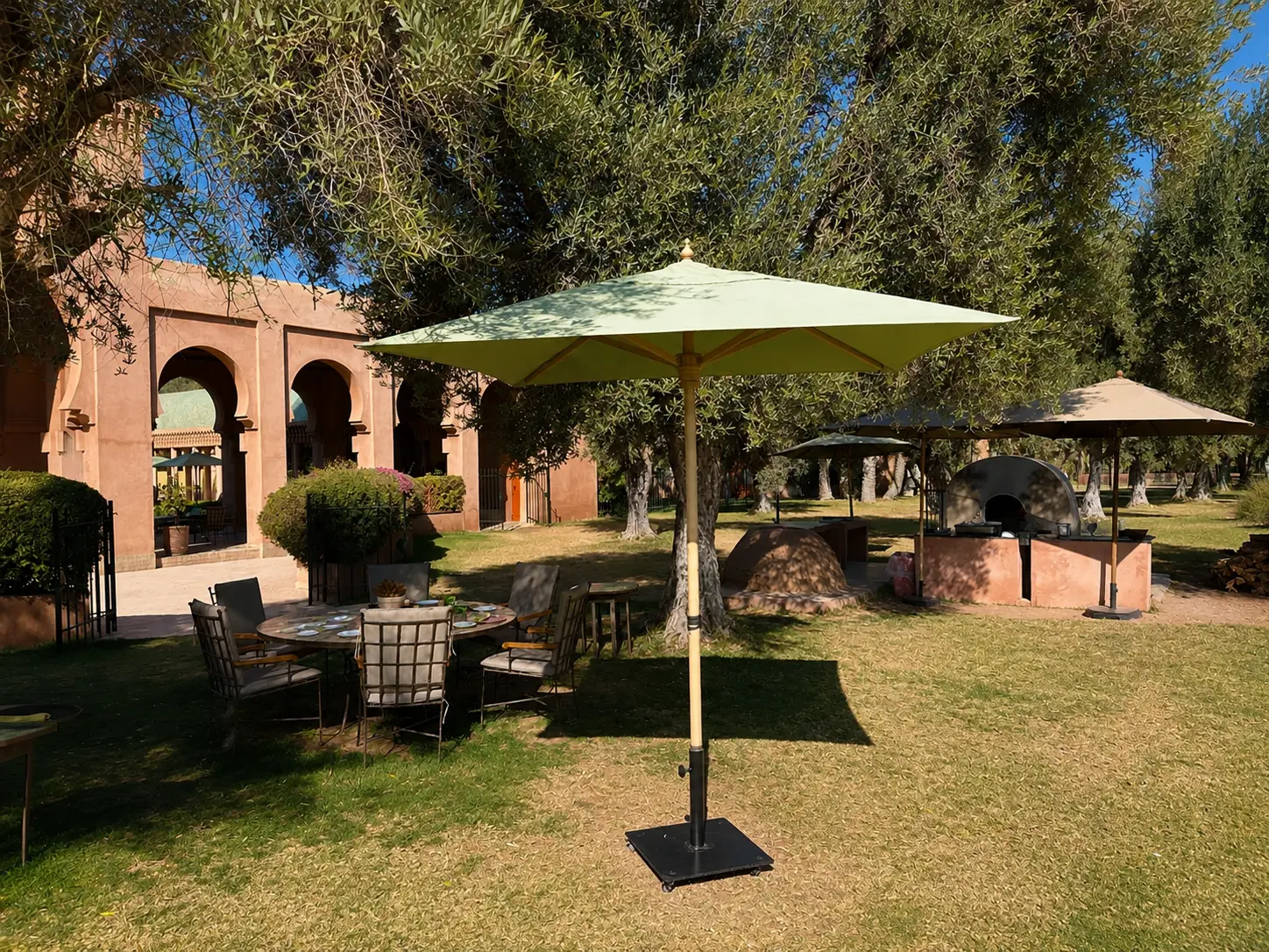 Outdoor lunch tables beneath mature olive trees at Amanjena's Olive Grove in Marrakech, with a wood-fired pizza oven and views across the resort gardens.