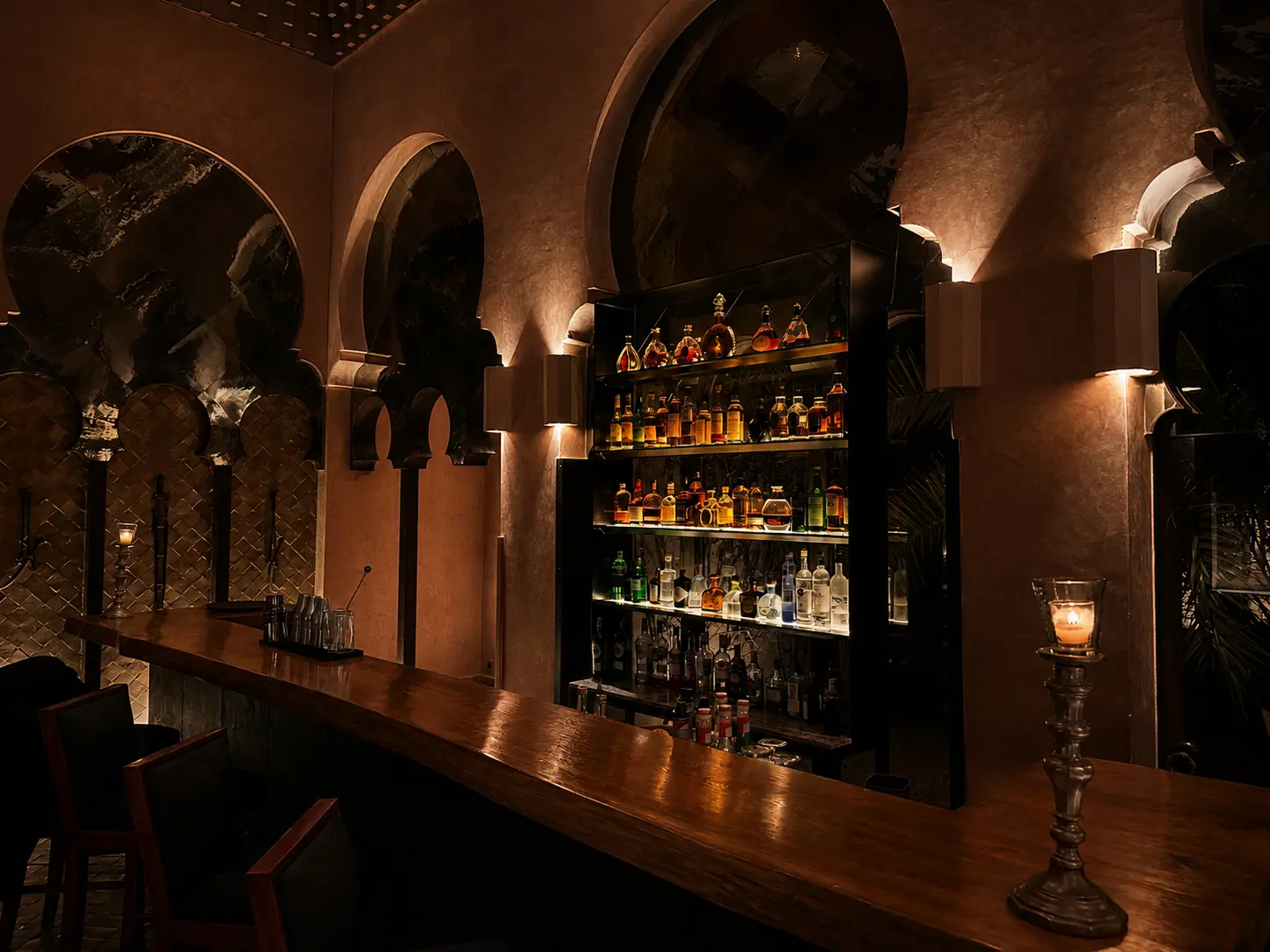 The candlelit bar at Amanjena in Marrakech, featuring Moroccan arches, a polished wood counter, illuminated spirits, and warm evening lighting.