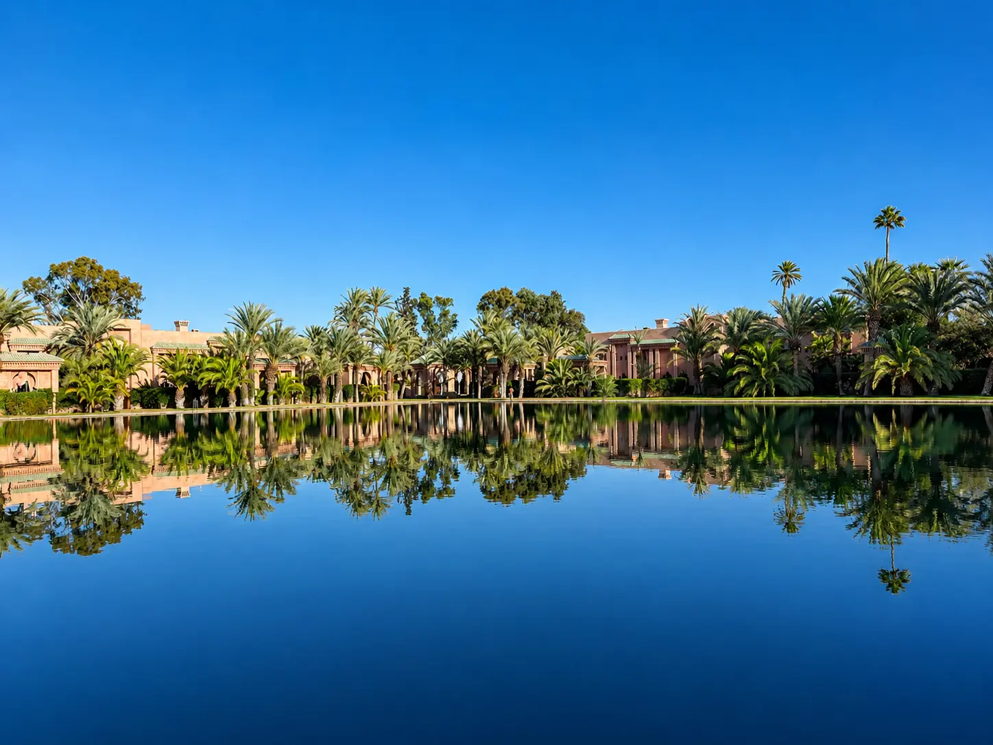 Amanjena’s main reflecting pool in Marrakech with palm trees, rose-colored pavilions, and Moroccan architecture mirrored across the water.