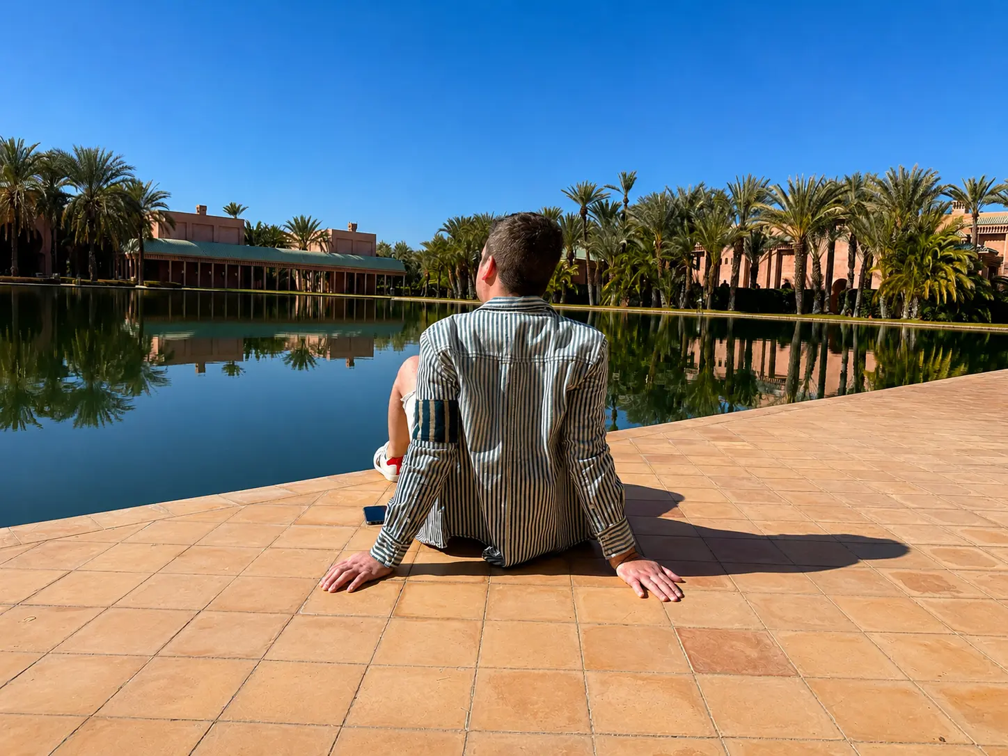 Guest sitting beside Amanjena’s main reflecting pool in Marrakech, surrounded by palm trees and rose-colored Moroccan architecture.