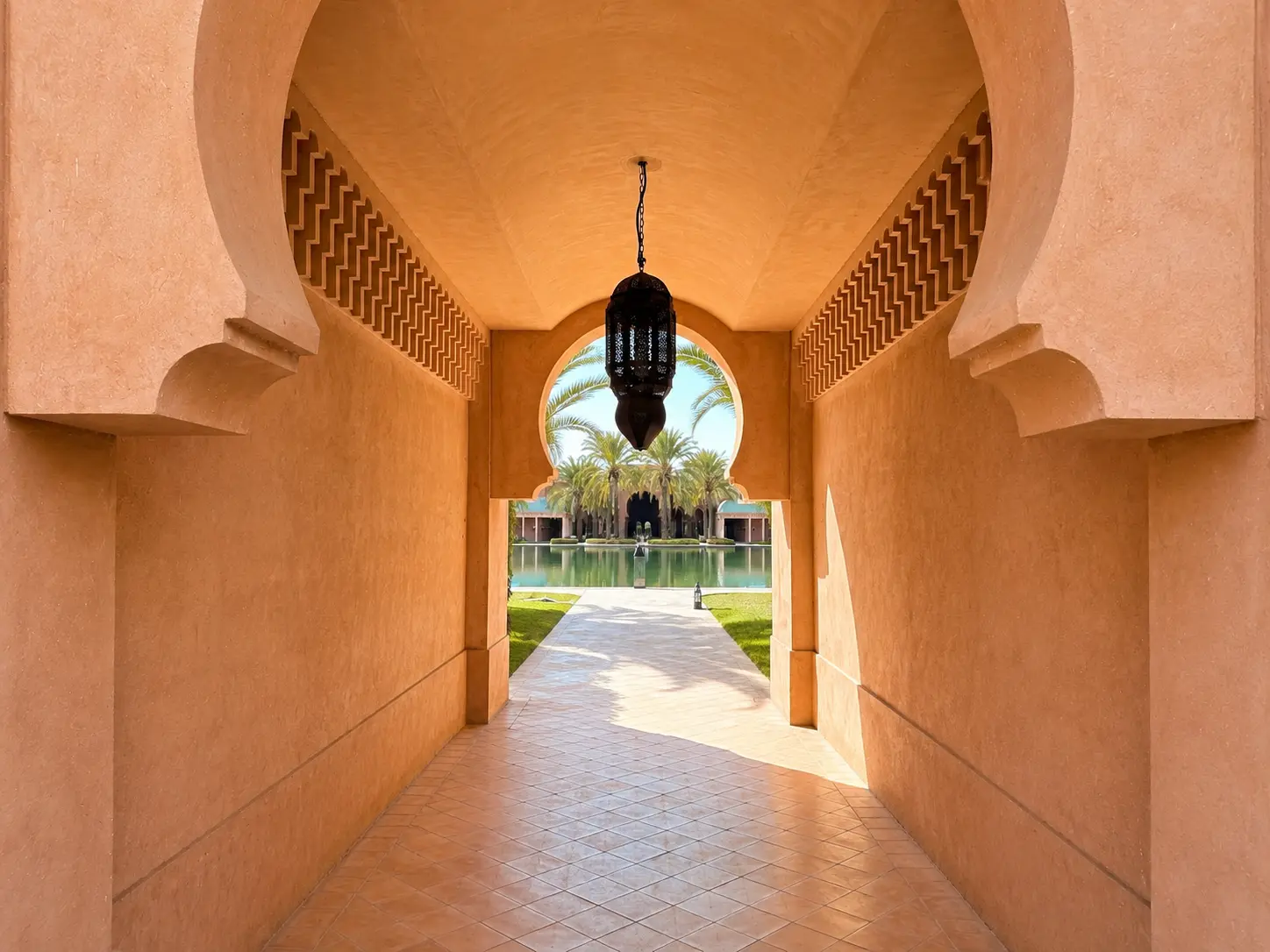 Symmetrical arched passage at Amanjena in Marrakech with a hanging Moroccan lantern and a view toward the resort’s main reflecting pool and palm-filled courtyard.